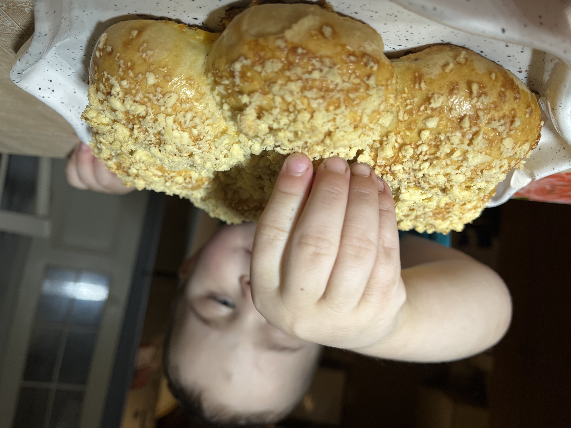 A child holds a bun or pastry with a crumb topping in their hand. The child has light skin and their hand is small with short fingernails. The background includes a blurry interior with a window and a dark corner. The location is Павловка, Russia.