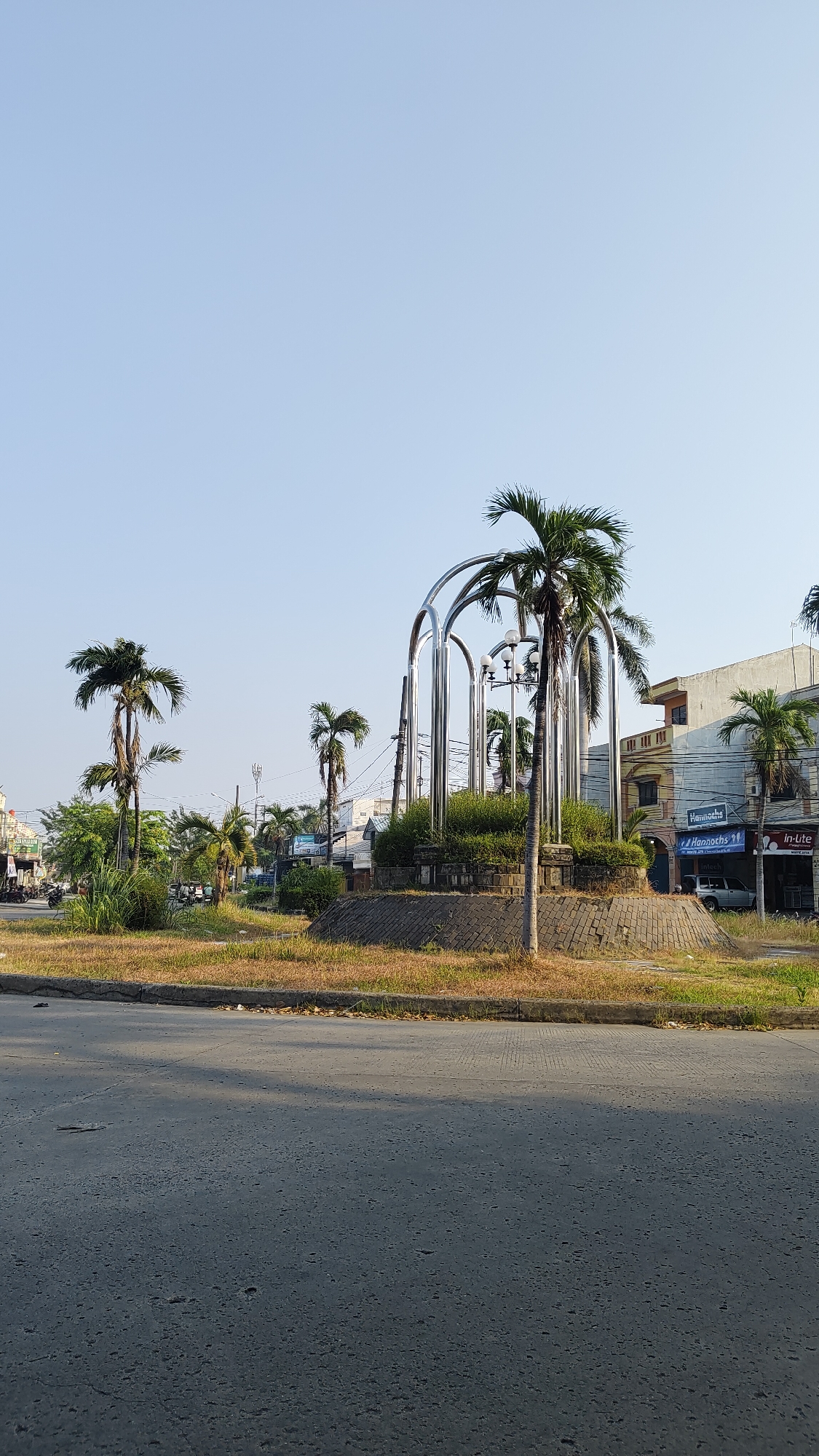The image depicts a street scene in Bekasi, Indonesia, featuring a circular monument with a metallic arch structure, surrounded by palm trees and various plants. The monument is situated on a traffic island, paved with brick. The background shows a row of buildings, some with signage indicating shops or businesses. A street with a few vehicles and motorbikes is visible to the left, suggesting moderate traffic. The sky is clear and blue, indicating a sunny day. The foreground shows a paved road.