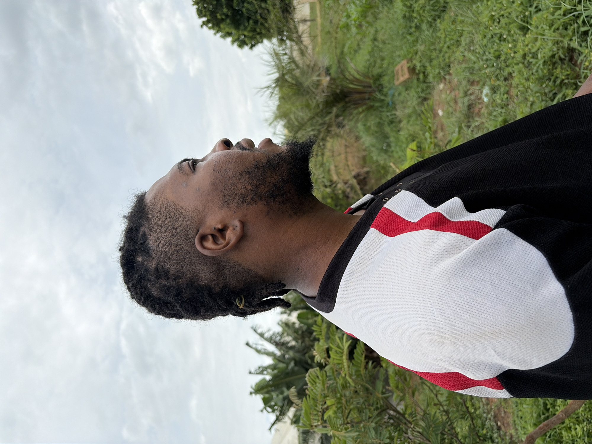 A medium close-up shot shows a Black male in profile, facing to the right, with his head tilted slightly upwards. He has dark skin, short dreadlocks styled with a faded undercut, and a goatee. He is wearing a black shirt with white and red paneling on the sleeves and chest. The background consists of lush green vegetation, including palm trees and leafy bushes, under an overcast sky. The setting appears to be outdoors, possibly in a rural or semi-urban area.