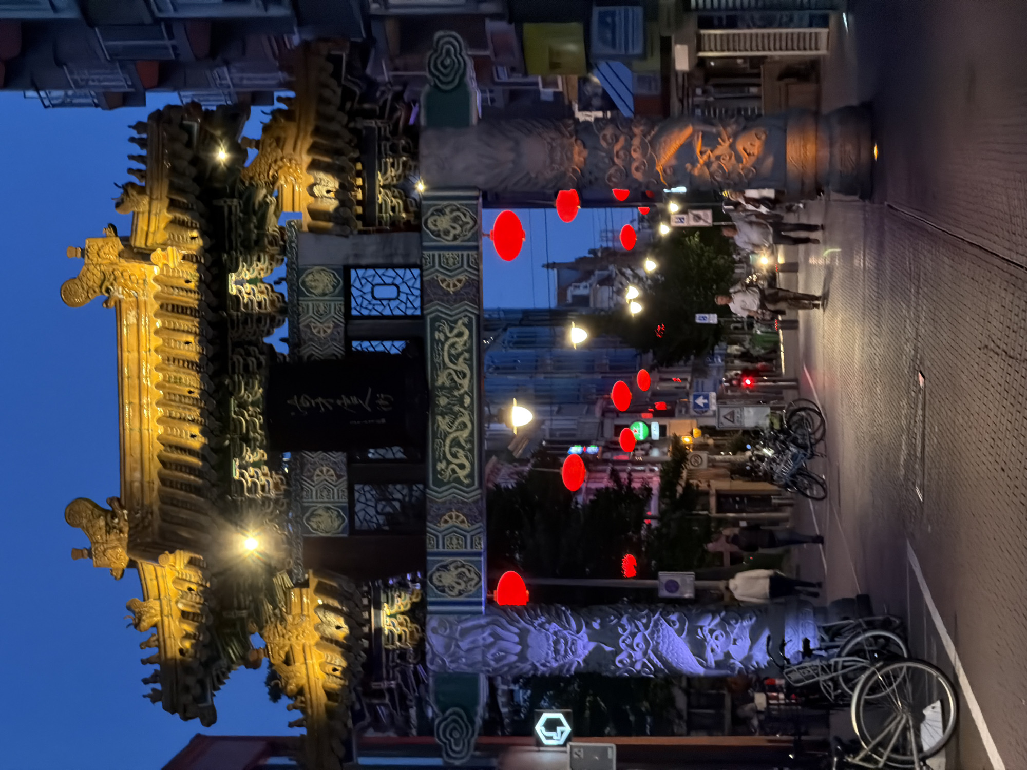 This image depicts the Chinatown Gate in The Hague, Netherlands, at dusk. The ornate, yellow-tiled roof of the gate is illuminated, casting a warm glow against the darkening blue sky. Red lanterns are strung across the street, adding vibrant pops of color. The gate itself is supported by large, stone pillars carved with intricate dragon motifs, which are lit with purple and orange lights.
In the foreground, several bicycles are parked along the street. The street is paved with brickwork. A few individuals are walking on the sidewalk and crossing the street. On the right side of the frame, buildings line the street, some with lit windows. Traffic lights and street signs are visible. The overall scene suggests a lively urban environment illuminated by artificial light as night falls.