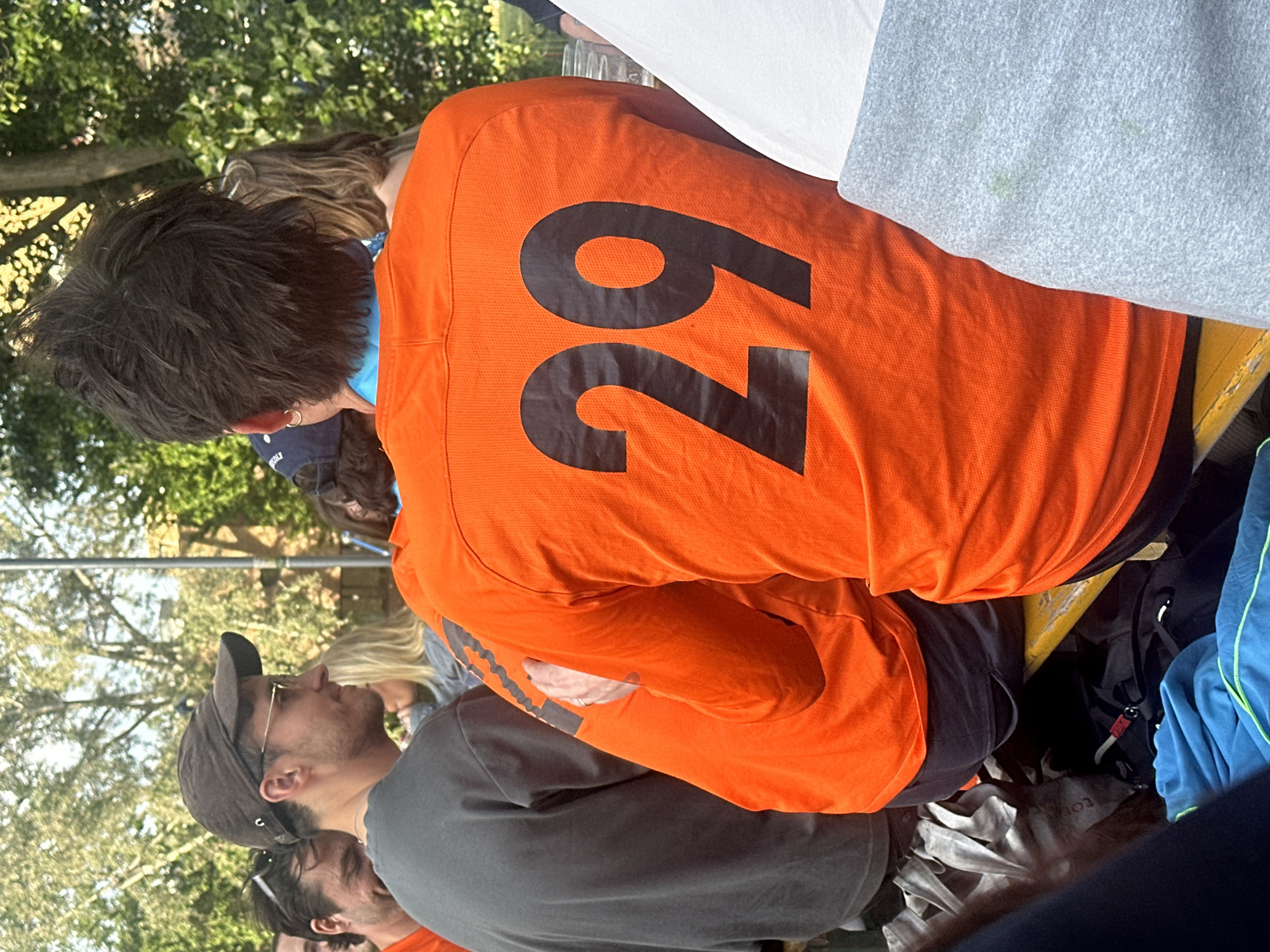 A slightly over-the-shoulder, eye-level shot shows a group of people sitting at a picnic table outdoors, likely on a sunny day. An individual wearing an orange shirt with a large black "29" on the back is in the foreground. Next to this person are other people; one is wearing a gray shirt, and another further down the table is wearing a baseball cap. Behind the "29" shirt, there are hints of more people. In the background, there are trees, indicating an outdoor park setting, and other indistinct figures. The context of the scene is unclear. It is not possible to determine if the location is in London, United Kingdom.