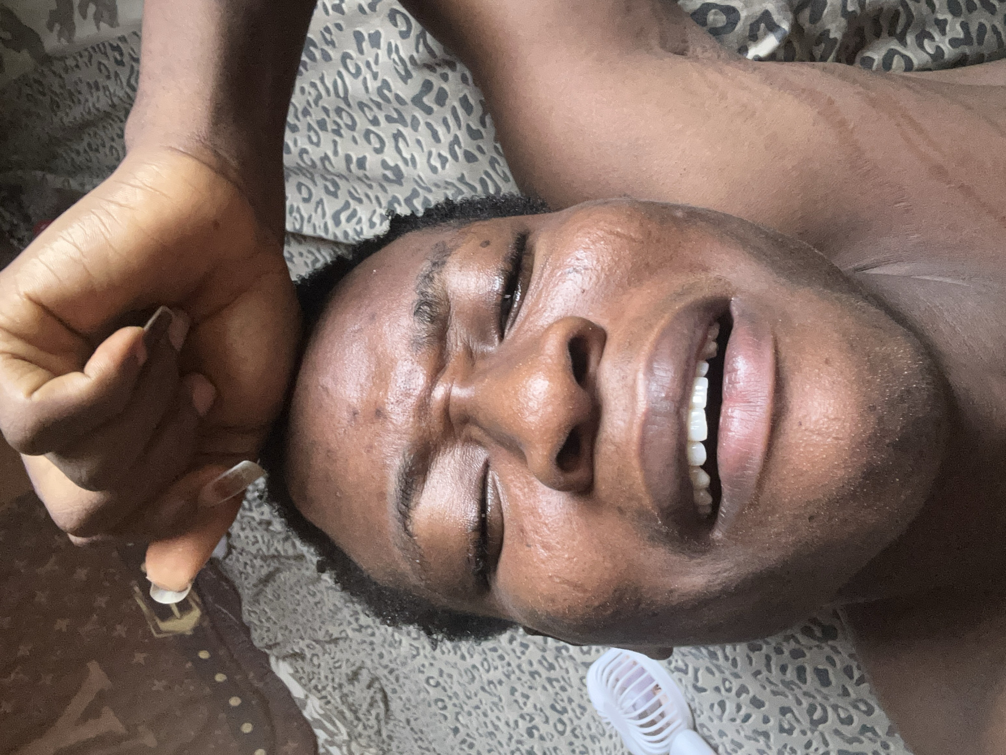 A close-up, portrait-oriented image features a dark-skinned individual lying on their back, with their eyes closed and a slight smile revealing their teeth. Their left hand is raised towards the top left corner of the frame, with long, white artificial nails visible. The person appears to be resting on a bed or surface covered with a tan and grey leopard-print fabric. Some stretch marks are visible on the person's chest. To the bottom right of the individual's face, a small, white plastic fan is visible.