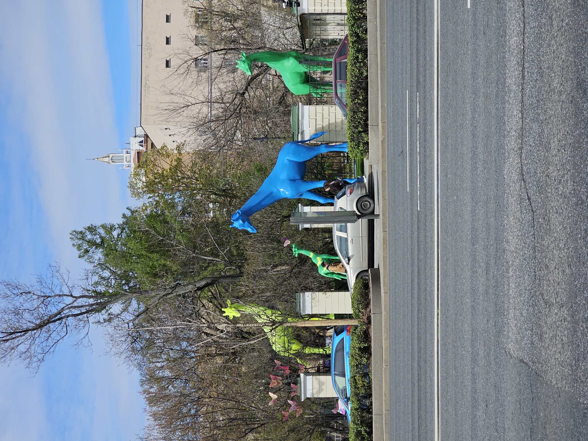 The image shows a street scene in Moscow, Russia, featuring colorful giraffe sculptures. There is a blue giraffe next to a white car parked along the curb. Further down the street, there are other giraffe sculptures in green. A blue car is also parked along the curb, further down the street near more green giraffe sculptures. The street has a grey asphalt surface with white lane markings. Trees and buildings are visible in the background, with a blue sky overhead. There is a building with a tower visible in the midground. The statues are separated from the sidewalk by a bush.