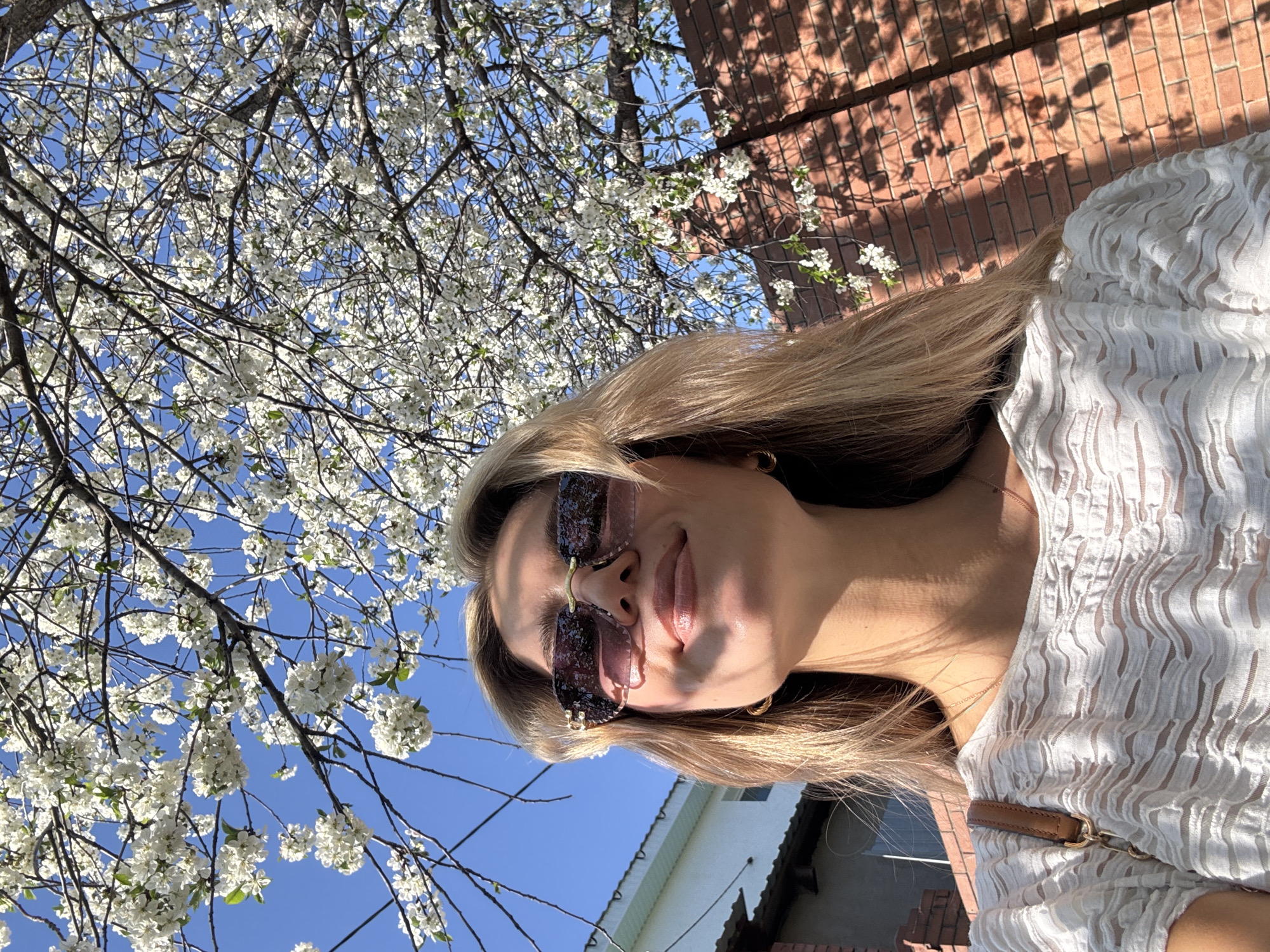 A woman stands beneath a blooming tree in an urban environment in the city of Unknown, Unknown. The woman has long, blonde hair, sunglasses, and is wearing a textured white top with a brown strap visible. She is positioned close to the camera, looking slightly down at it. Overhead, a tree branches out, covered in white flowers against a clear blue sky. In the background, there is a brick wall with tree shadows and a building with a white wall and roof.