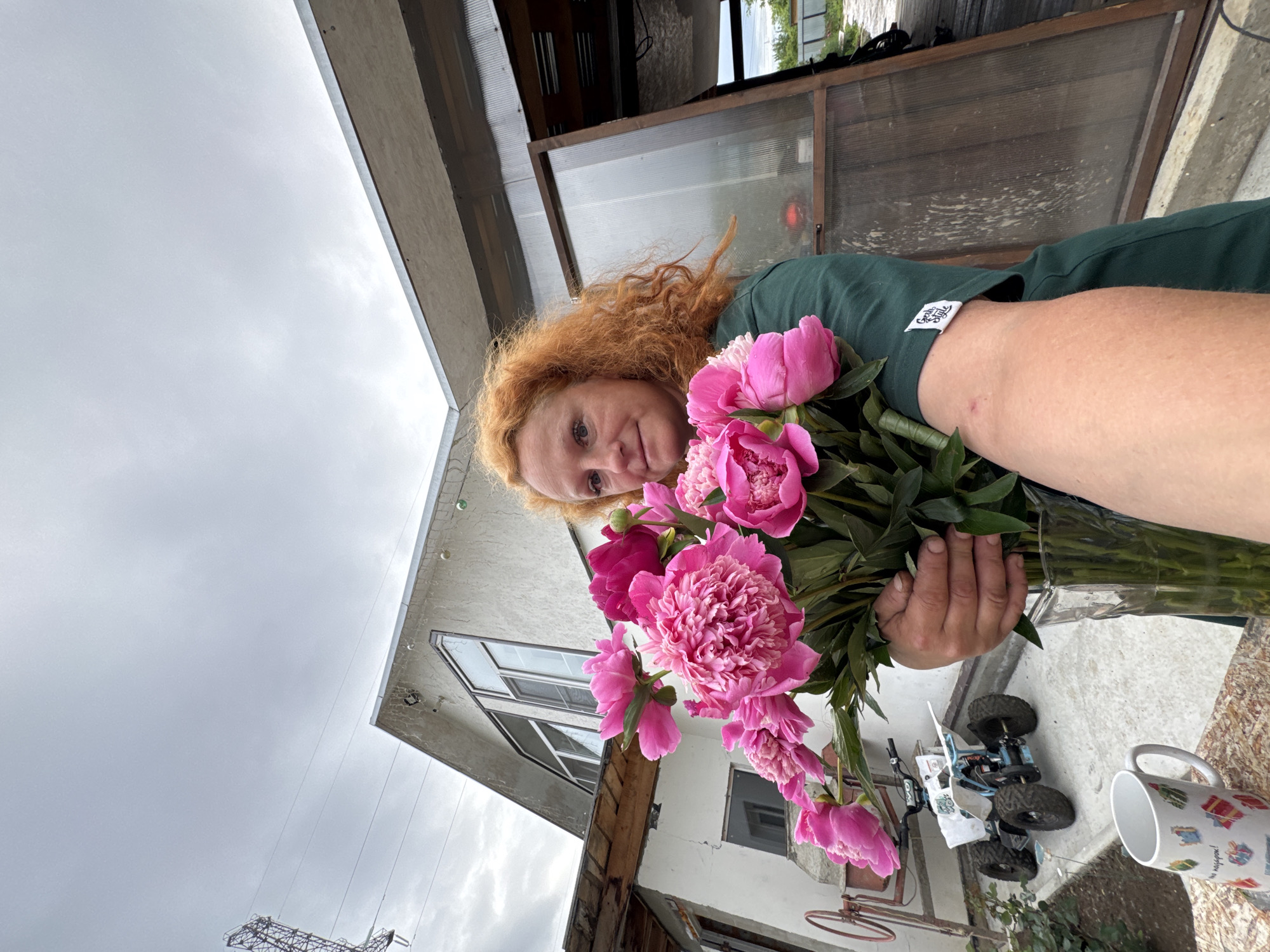 In an outdoor, overcast setting, a person with vibrant red, curly hair holds a large bouquet of bright pink peonies. They are wearing a dark green t-shirt with a small white tag on the sleeve. The background features a white, stucco-style building with angled windows, suggesting a residential or rural property. To the right, a greenhouse-like structure with translucent walls and a wooden frame is visible, with its door ajar. In the foreground, on the left, a white mug with colorful drawings sits on a surface, and a small ATV-like vehicle is parked on a paved area. The overall atmosphere is casual and suggestive of a home or garden environment. This scene is likely located in the city of Chaikovsky, Russia, given the details of the architecture and the commonality of such outdoor scenes in rural and suburban areas of the country.