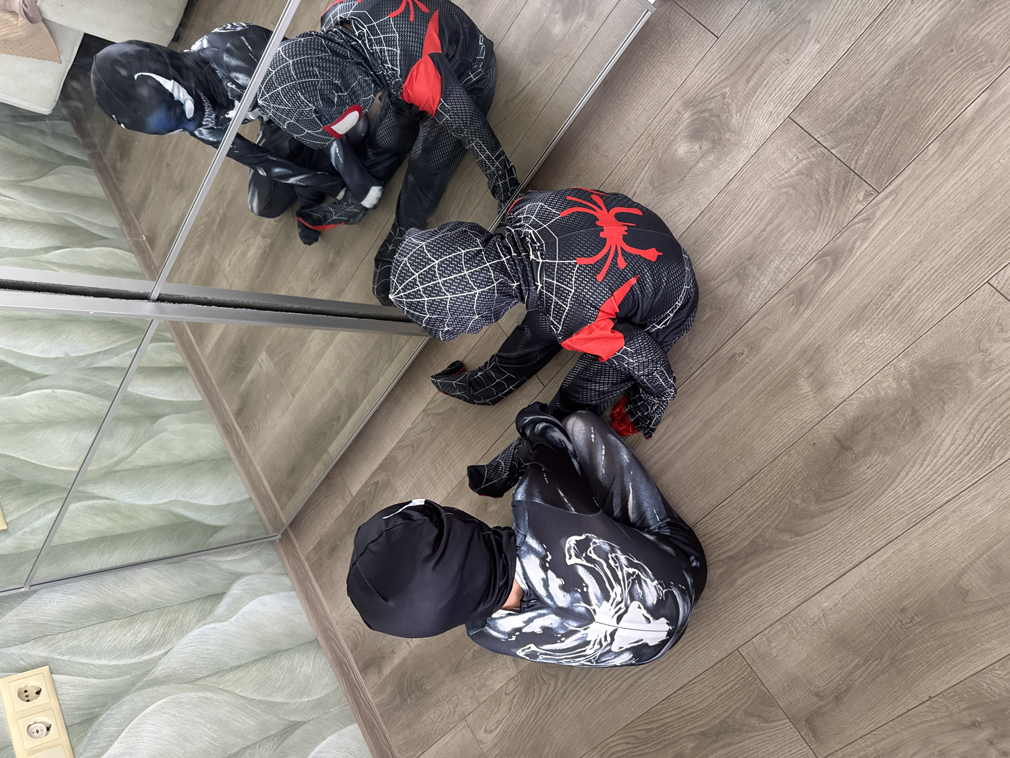Two children dressed in superhero costumes are sitting on a wooden floor in front of a large mirror. The child on the left is wearing a Venom costume with a black hood and a white, skeletal-like print on the suit. The child on the right is wearing a Spider-Man costume with red accents and a spider web pattern. The mirror reflects the children and the room, showing another child in a Venom costume and another in a Spider-Man costume. The wall behind them is decorated with a light green patterned wallpaper. Electrical outlets are visible on the wall near the top left corner. The scene appears to be indoors, possibly in a bedroom or playroom. The location is Sovkhoza imeni Lenina, Russia.