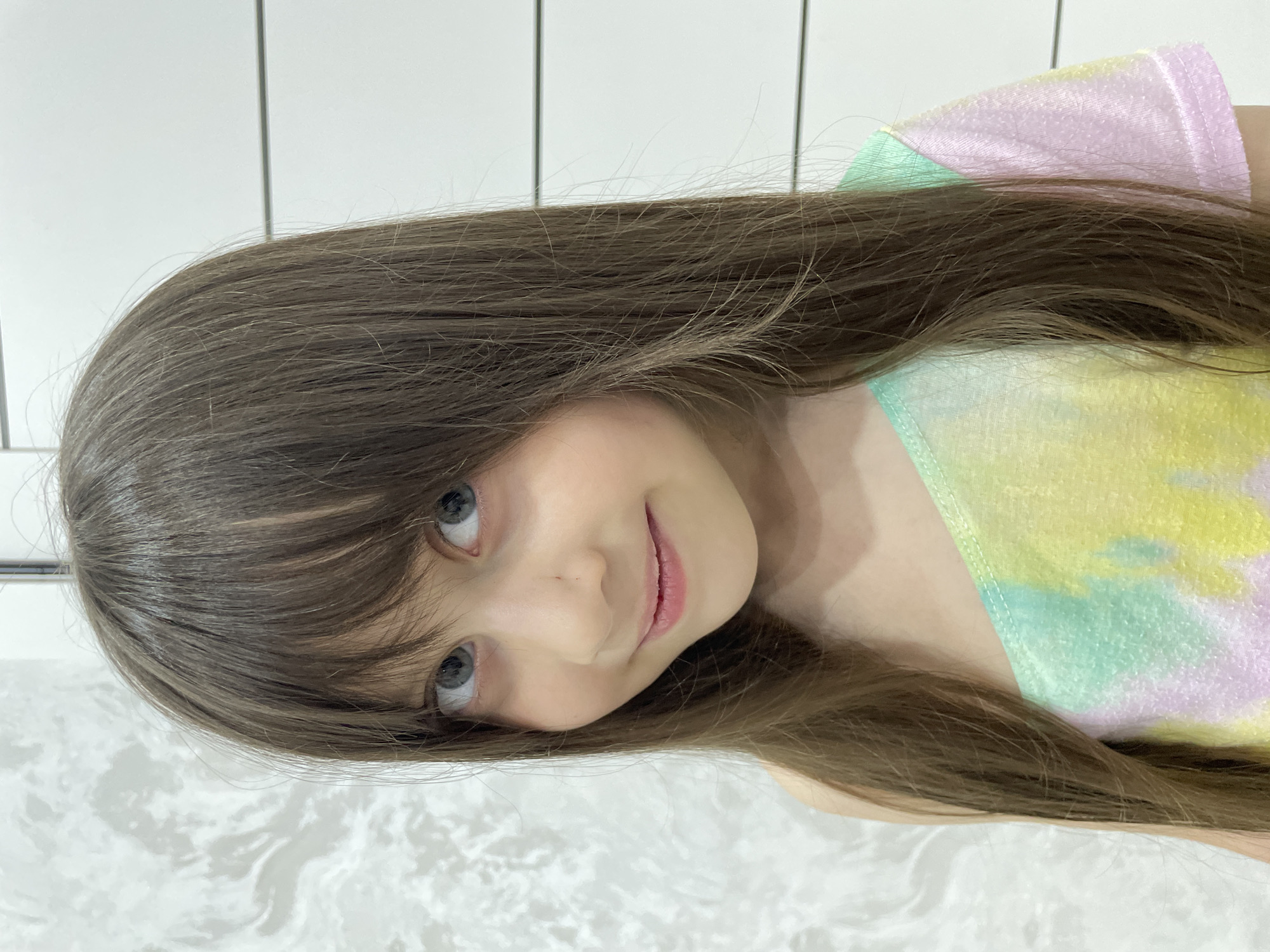 A young girl with long, brown hair and blue eyes is facing the camera with a slight smile. She is wearing a pastel-colored tie-dye shirt with pink, yellow, and mint green hues. The background consists of a white tiled wall and a white marble-like surface. The image is rotated ninety degrees counter-clockwise. There is no information in the image to suggest it was taken in Izhevsk, Russia.