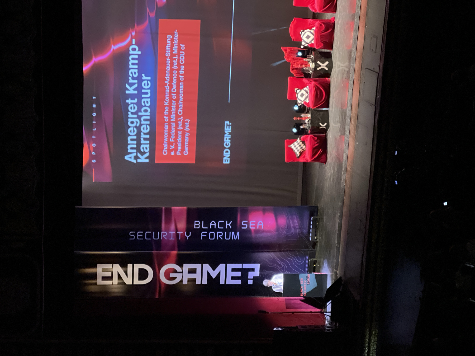 The image shows a stage setting for the "Black Sea Security Forum", likely taken at a location in Odesa, Ukraine, given the event's theme. The stage features a backdrop with the forum's name and the question "END GAME?". Adjacent to the main backdrop is a screen that displays the name "Annegret Kramp-Karrenbauer" along with her titles: "Chairwoman of the Konrad-Adenauer-Stiftung e. V., Federal Minister of Defence (ret.), Minister-President (ret.), Chairwoman of the CDU of Germany (ret.)". A series of red velvet chairs, each accompanied by a small table displaying an "X" symbol, are arranged on the stage, presumably for panelists or speakers. To the right of the chairs is a dark area with what appears to be part of the stage extending into the audience. At the center stage is a lectern and a microphone. The image is a photograph taken from the audience facing the stage.