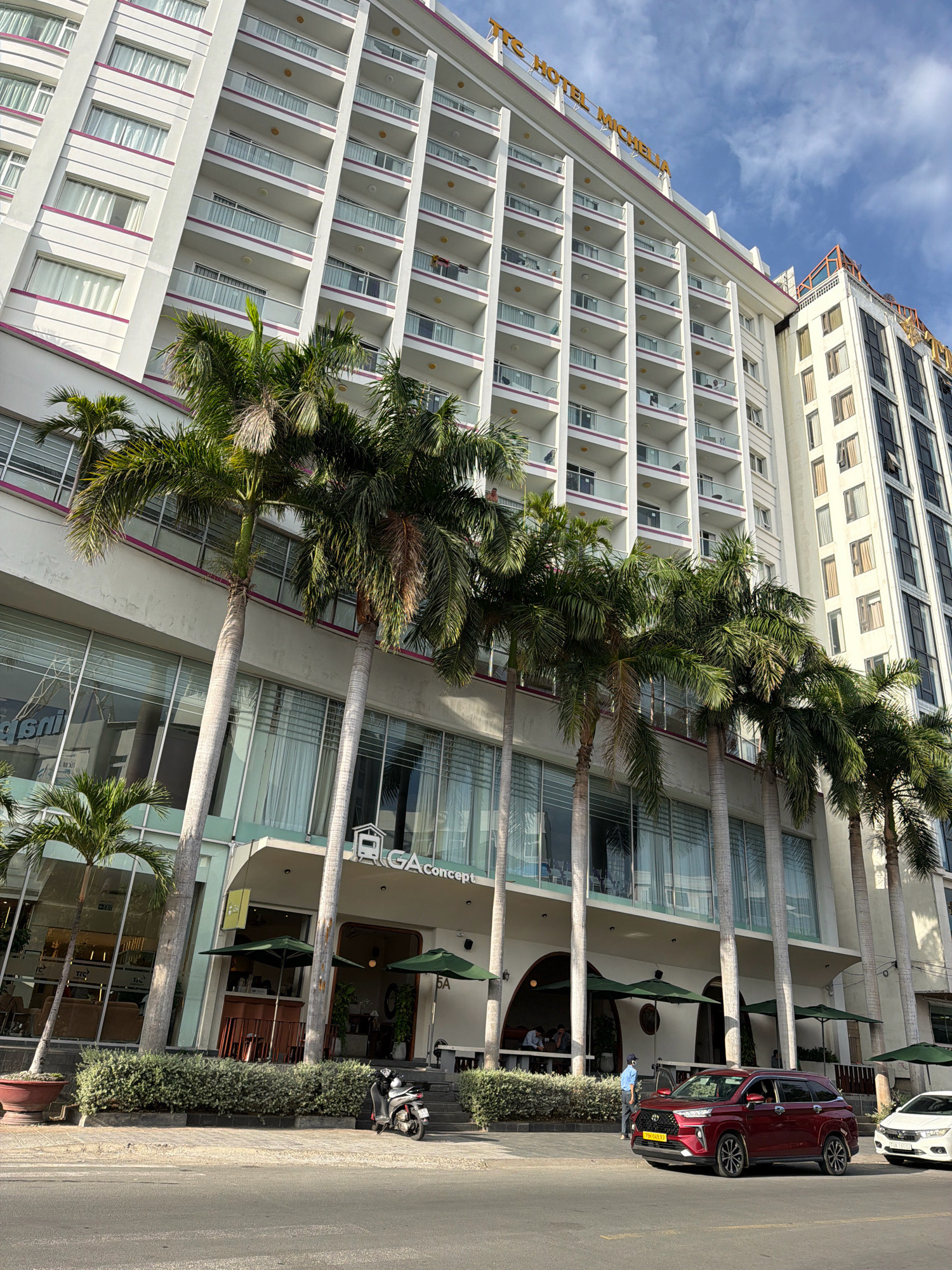 The image shows a street scene in Nha Trang, Vietnam, focusing on a large, modern hotel with a white facade and pink trim, identified by the signage "TTC HOTEL MICHELLIA" at the top. Palm trees line the front of the hotel, some partially obscuring the lower levels, which feature large glass windows and an entrance area with awnings. A dark red SUV is parked in front of the hotel, with its driver's side door open and a person standing beside it. A white car is parked to the right of the SUV. A scooter is parked on the sidewalk in front of a section of dense green bushes. The sky is partly cloudy.