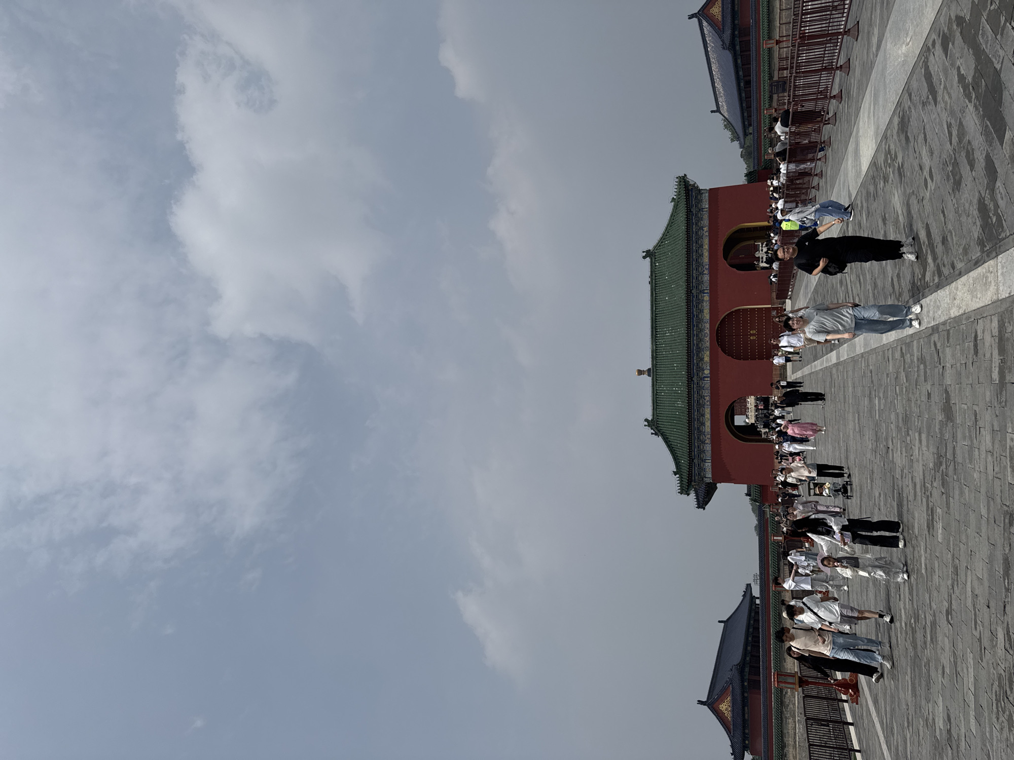 The image depicts a courtyard scene in the Dongcheng District, China. A red building with a green roof is prominent on the right, with arched doorways. People are walking across the stone-paved courtyard. The sky is cloudy and overcast. Additional buildings with upturned roof corners and architectural details flank the main structure. A fence is visible.