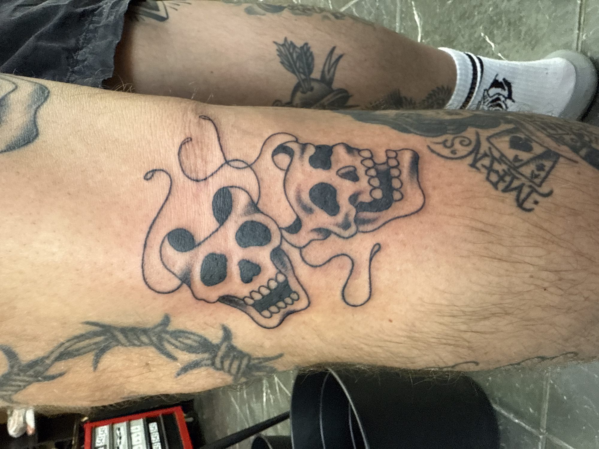 The image shows a person's leg with several tattoos. The most prominent tattoo features two melting skulls connected by strands of what appears to be melting matter. Additional tattoos include barbed wire, a playing card design, and a bird with an arrow through it on the upper leg. The person is wearing a sock with a logo on it. The leg is positioned against a light-colored, possibly tiled, surface with a black object in the foreground. There is no indication of Moscow, Russia.