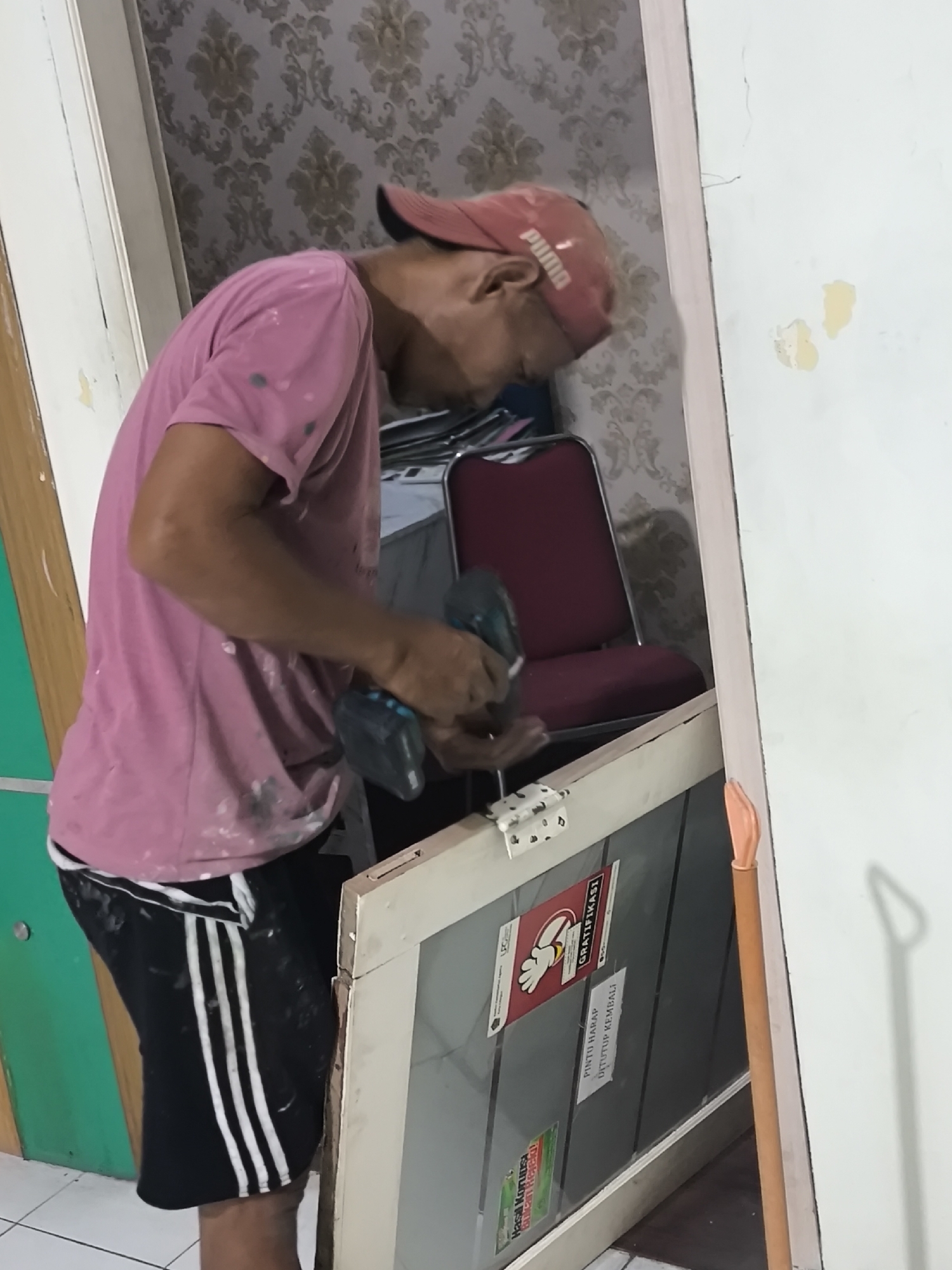 A man wearing a pink t-shirt and a red baseball cap is seen in Cilegon, Indonesia, working on a door. He is holding a cordless drill and appears to be in the process of attaching or adjusting hinges. The room has patterned wallpaper and a red chair is visible in the background. The man's clothing is stained, suggesting he is involved in a construction or renovation project. A partially assembled door frame and the door itself are the central focus of his activity.