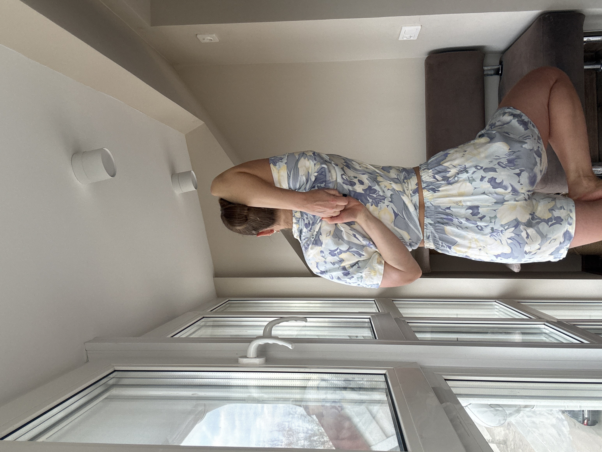 A person is standing in a room with a large window. The person is wearing a floral print pajama set. The room appears to be inside a building, and there are objects like a couch visible. The location appears to be in the city of Chaikovsky, Russia.