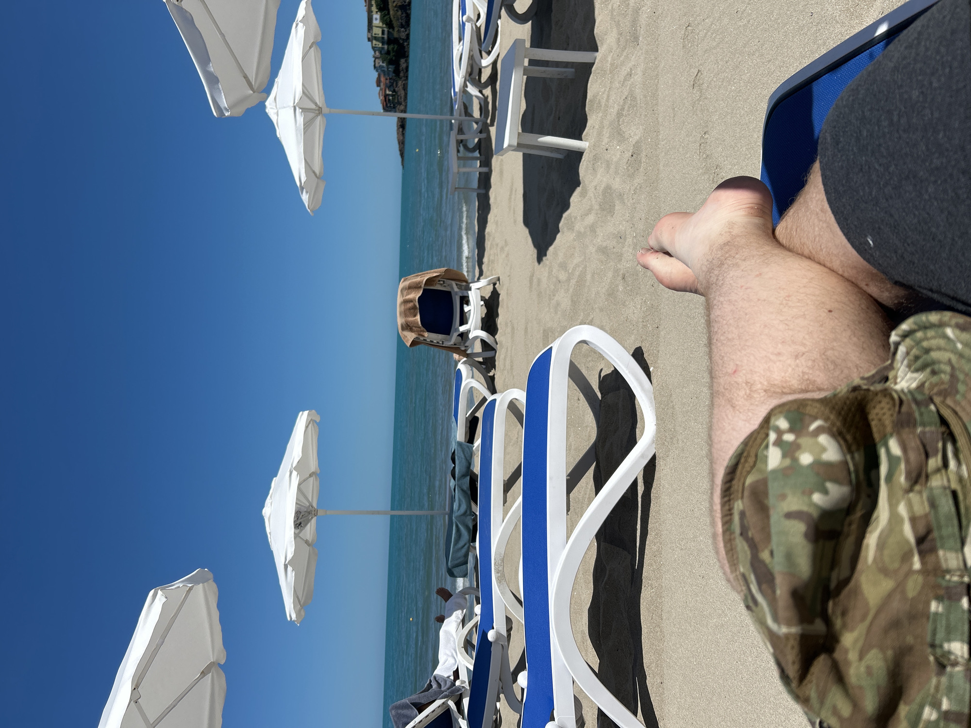 This is a first-person perspective photo taken from a lounge chair on a sandy beach in Sozopol, Bulgaria. The person is wearing camouflage shorts and dark grey sweatpants. Their legs are crossed, and their bare feet are visible. In the foreground, two white lounge chairs with blue cushions are partially visible. Beyond these chairs, more lounge chairs and white umbrellas are scattered across the sand. The calm, turquoise sea stretches to the horizon, under a clear blue sky. In the distance, buildings are visible on a rocky cliffside, characteristic of Sozopol's coastline. The scene suggests a relaxed day at the beach.