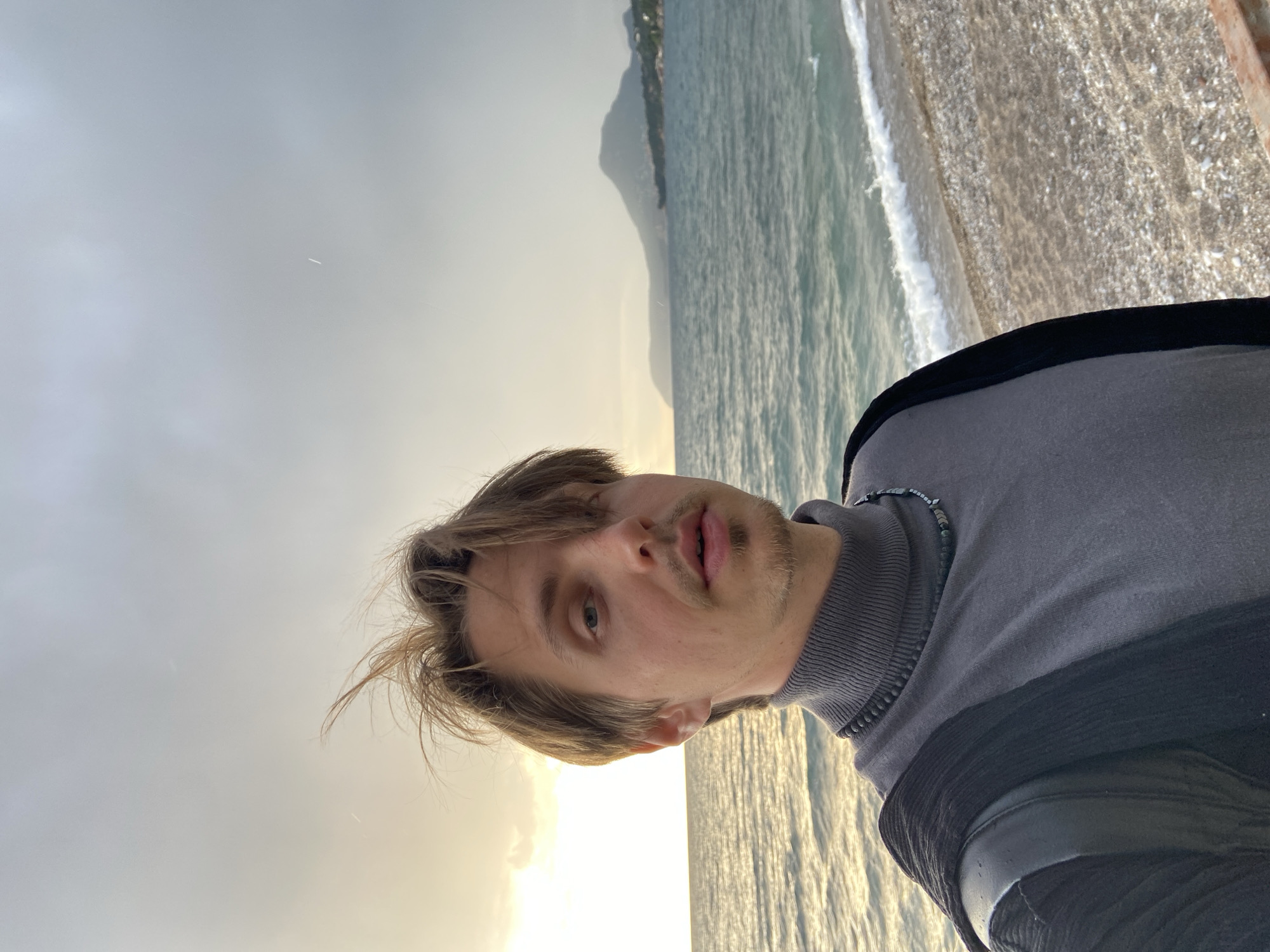 A young man with light brown, medium-length hair and light stubble is depicted in a selfie, looking directly at the camera with a neutral expression. He is wearing a grey turtleneck sweater, under which a dark, possibly beaded, necklace is visible. He also appears to be wearing a dark, textured outer layer, possibly a jacket or vest.
The scene captures a coastal environment. To the right of the man, the sea extends, reflecting a warm, golden light on its surface. Further to the right, a pebbly or rocky beach is visible, where gentle waves are breaking. In the distance, across the water, a range of mountains or hills can be seen under a hazy, partially cloudy sky. The lighting, characterized by the golden glow near the horizon, suggests it is late afternoon or early evening, likely near sunset. The overall weather appears overcast but brightened by the low sun. The location is a seaside setting, consistent with Bar, Montenegro. No text is visible in the image.