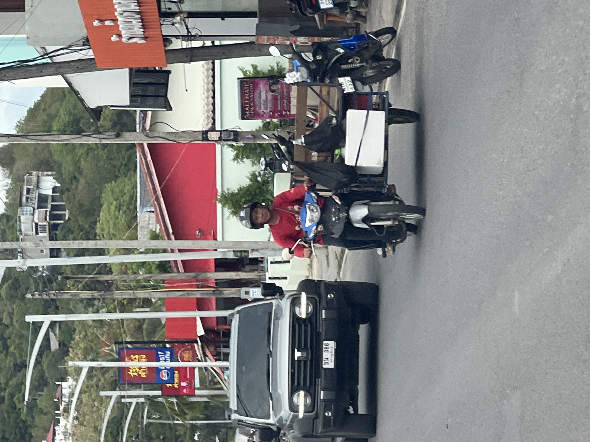 In บ้านบางรักษ์, Thailand, on a paved road with visible lane markings, a silver SUV is seen in the foreground. To the right of the SUV, a man wearing a red long-sleeved shirt and a black helmet rides a white and blue motorcycle with a sidecar. The sidecar is carrying a white cooler and two black bags. In the background, there are buildings with red roofs and white facades, electric poles with wires, and a tree-covered hill. A red and orange sign is visible on the right side of the image.