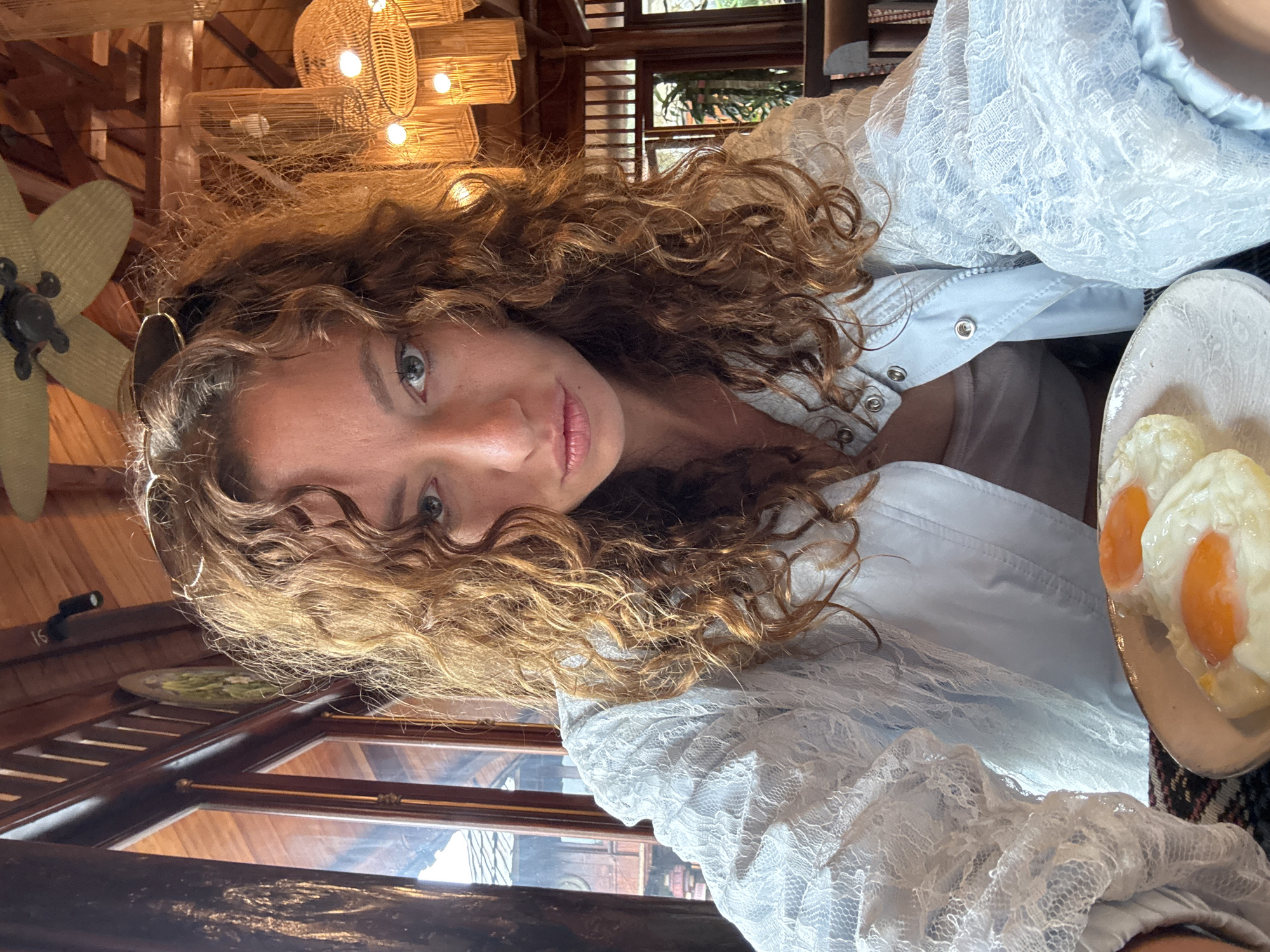 A woman with curly auburn hair and light blue eyes looks at the camera with a pout. She is wearing a light blue lace jacket over a light colored tank top and has sunglasses on her head. In front of her is a plate with two fried eggs. She is seated at a table in a restaurant with wooden accents and rattan light fixtures. The scene is likely Moc Chau, Vietnam, known for its agricultural landscapes and tea plantations.