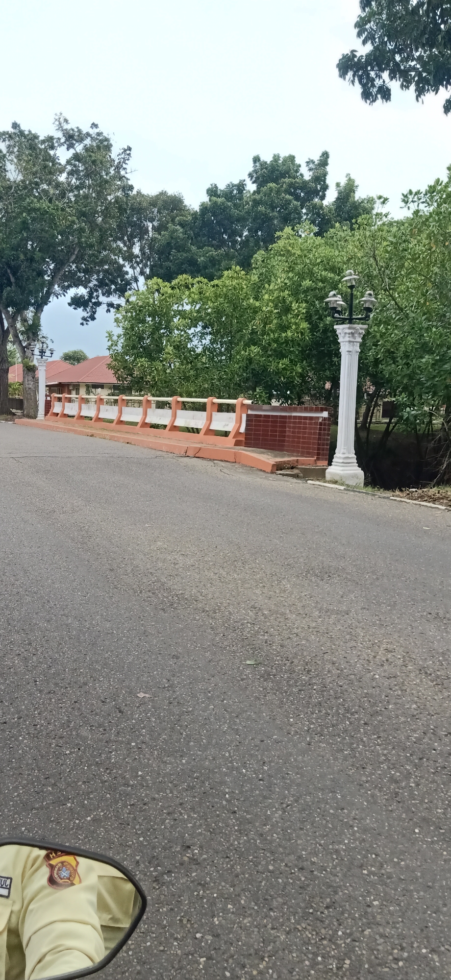 The image shows a view from a road, possibly on a motorcycle, as indicated by the reflection of a person in a uniform in the lower left corner. The road surface is asphalt. In the mid-ground, there is a bridge or elevated walkway with an orange and white railing. To the right of the walkway, there is a decorative white lamp post with multiple lights. Behind the walkway and lamp post, there is a dense green foliage of trees and some buildings with red tiled roofs. The sky is overcast. The location is described as Tambontunong, Indonesia.