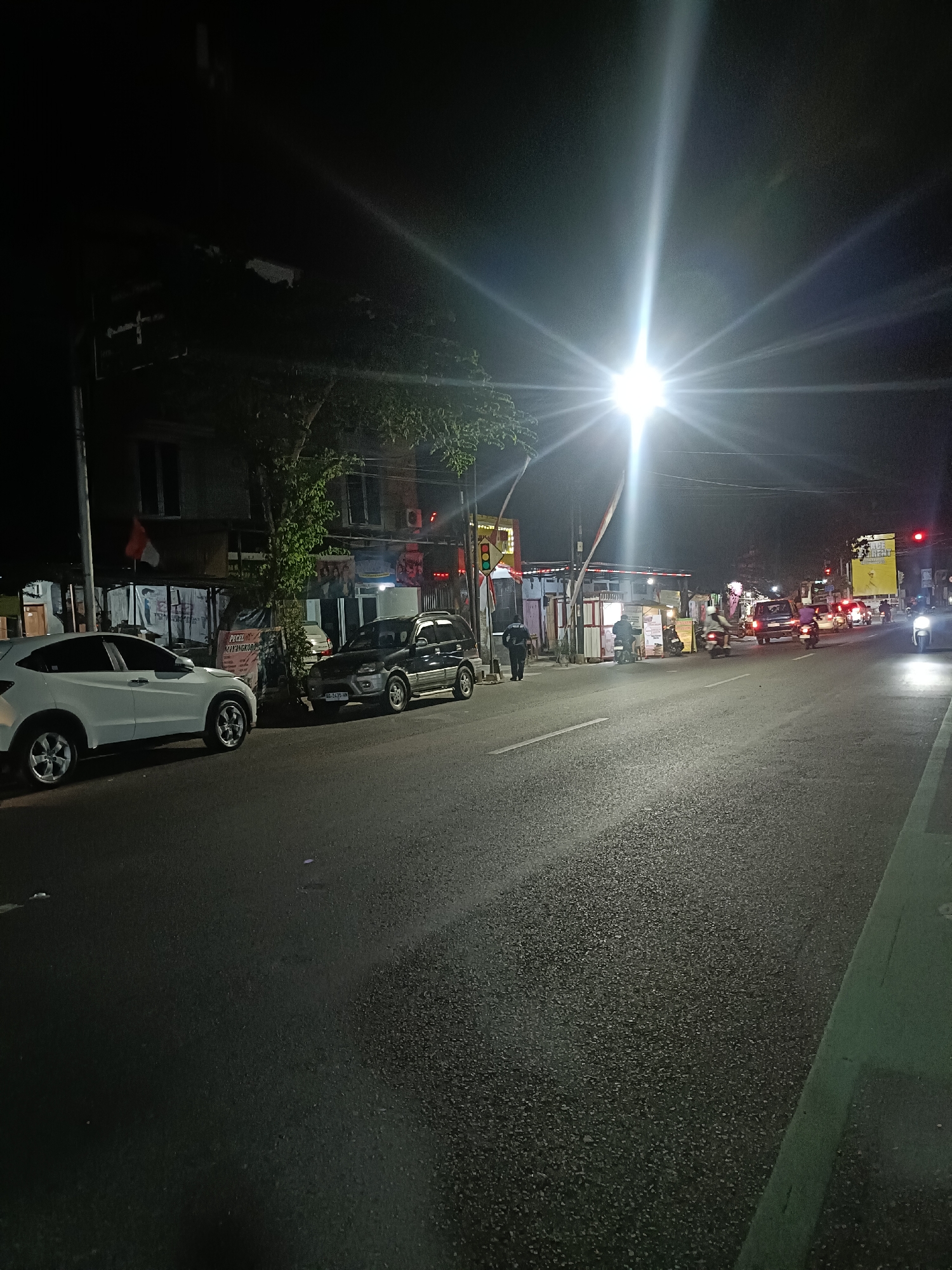 A nighttime scene on a street in the city of Kediri, Indonesia, features a bright street lamp illuminating the asphalt road. Several vehicles, including cars and motorcycles, are present on the road, some of which appear to be in motion. Buildings line the side of the street, with a few displaying illuminated signs. One building has a red and white flag displayed. The overall atmosphere is one of a moderately busy urban environment at night, with artificial light sources dominating the visual field.