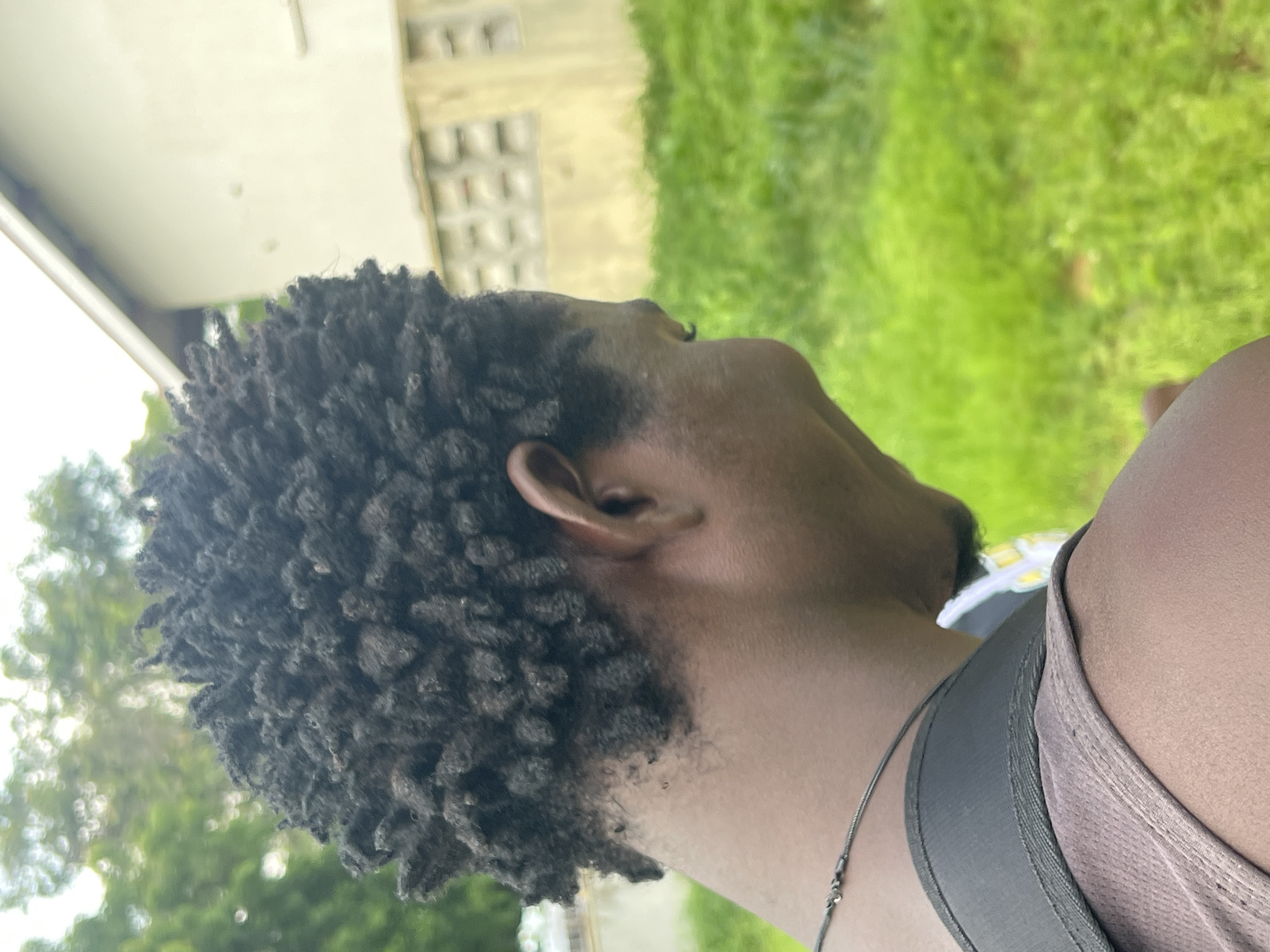 A medium shot shows the profile of a person from the neck up. The person has dark, short dreadlocks and is looking down and to the right. They are wearing a brown tank top and a black backpack strap is visible over their shoulder. A thin silver necklace is also visible. The background is out of focus and shows a building with light-colored walls and some greenery, likely in an outdoor setting in the city of Jalingo, Nigeria. The overall lighting is bright and natural.