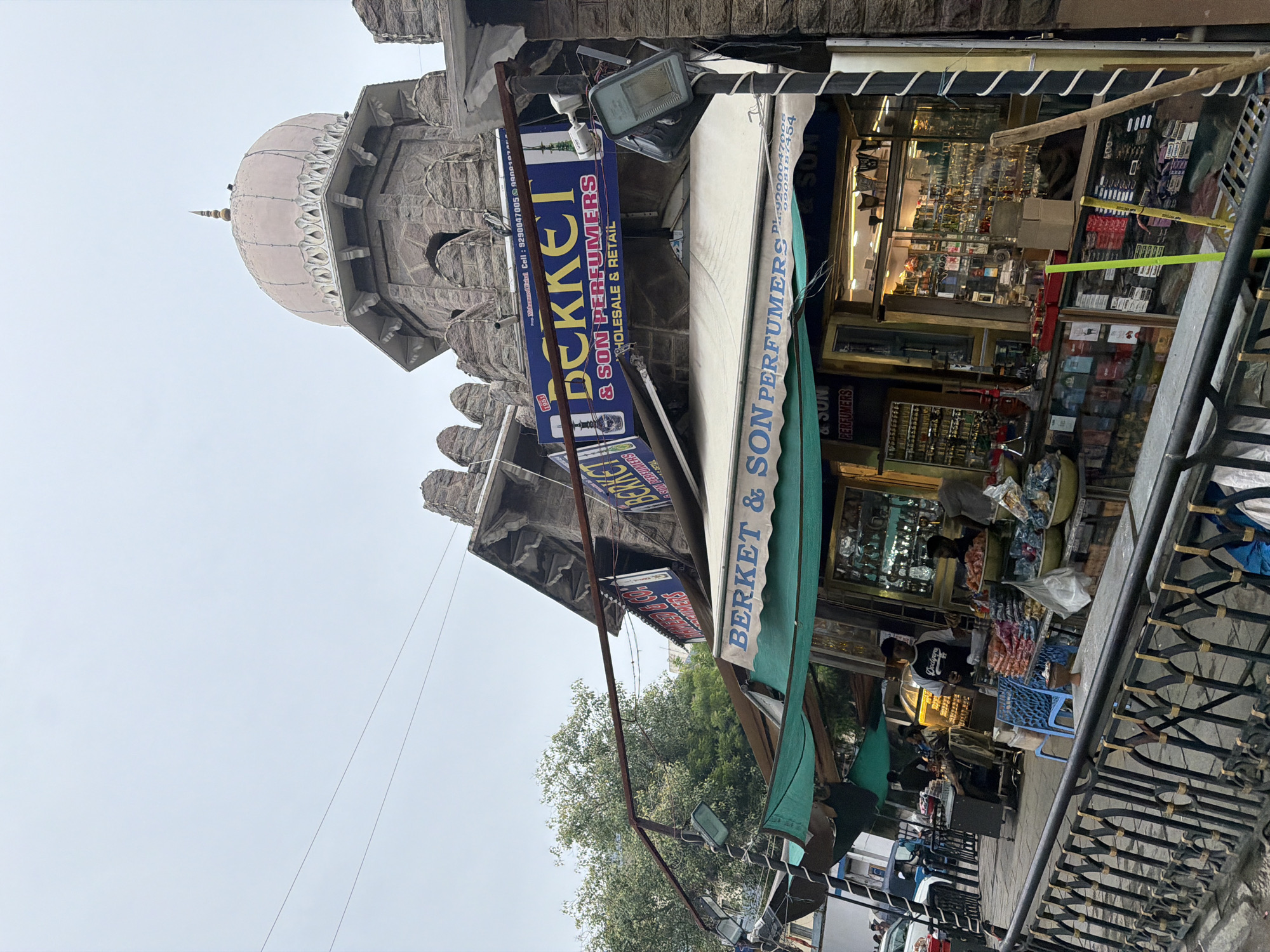 This is a vertically oriented photograph of a street scene in Hyderabad, India. The image captures a storefront displaying perfumes and other retail items. A prominent sign reads "BERKET & SON PERFUMERS WHOLESALE & RETAIL," with a phone number also visible. The store's interior is filled with shelves of products, visible through a glass storefront.
In the background, a stone structure with a dome, possibly a historical building or a temple, is partially visible. The sky is overcast, suggesting a cloudy day. Several people are seen in and around the shop, with some interacting with merchandise and others standing. One person appears to be a shopkeeper attending to customers. The overall impression is of a bustling marketplace. The time of day appears to be daytime.