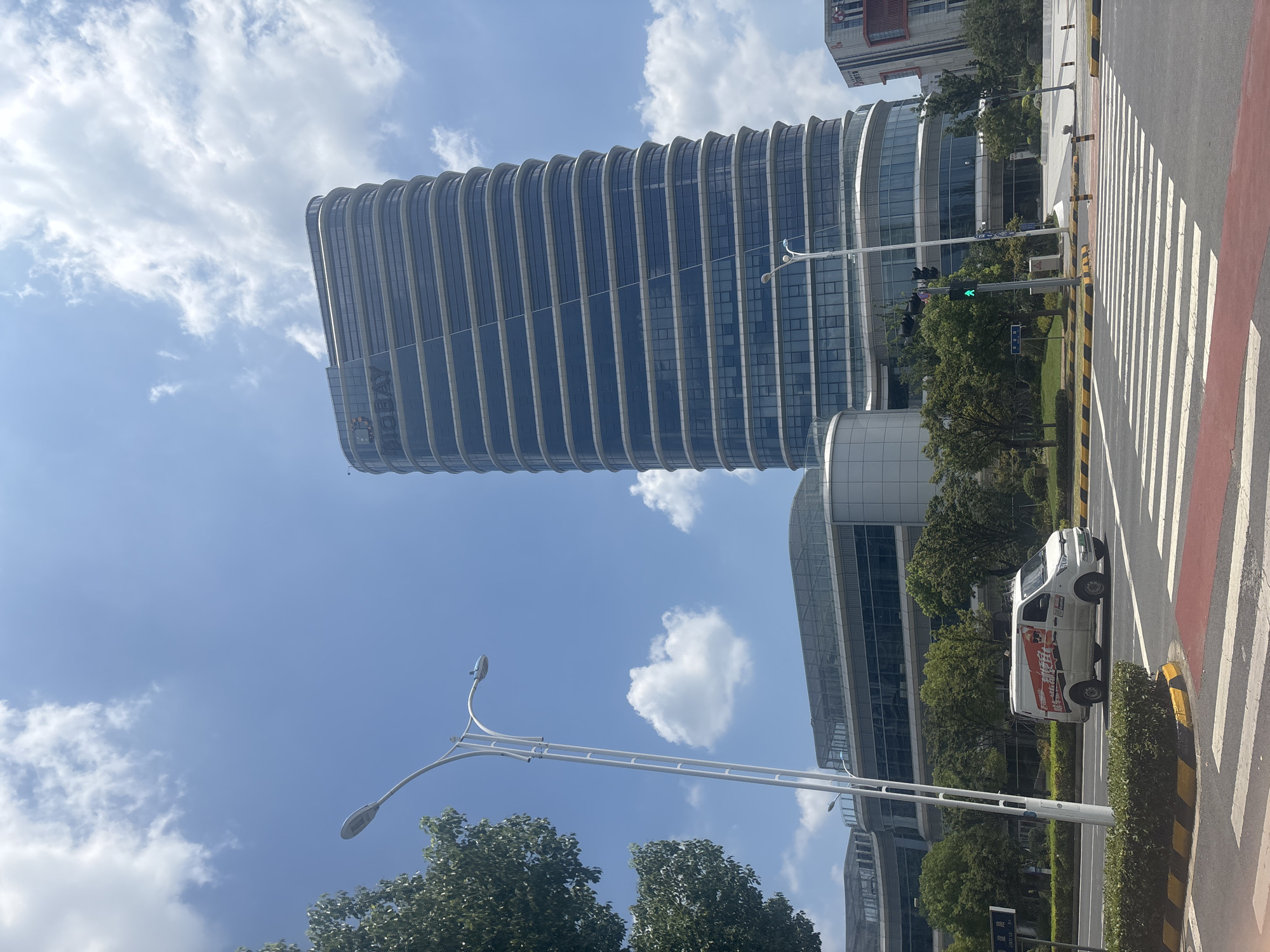 The image depicts a view of the BioBAY (Biotechnology and Pharmaceutical Industry Park) in Suzhou Industrial Park, China. In the foreground, a white delivery van with Chinese characters and graphics on its side is driving across a pedestrian crossing. The van is positioned on the right side of the road. A lamppost stands on the left side of the road, and behind it, a large, modern, multi-story building with a curved facade of glass and metal panels dominates the background. The building features horizontal banding. To the left of this main building, another, lower building with a glass facade is visible. Trees and green spaces are present around the buildings and along the roadside. The sky is blue with scattered white clouds. A traffic light displaying green is visible on the lamppost. The overall scene suggests an urban environment with commercial and industrial activity.