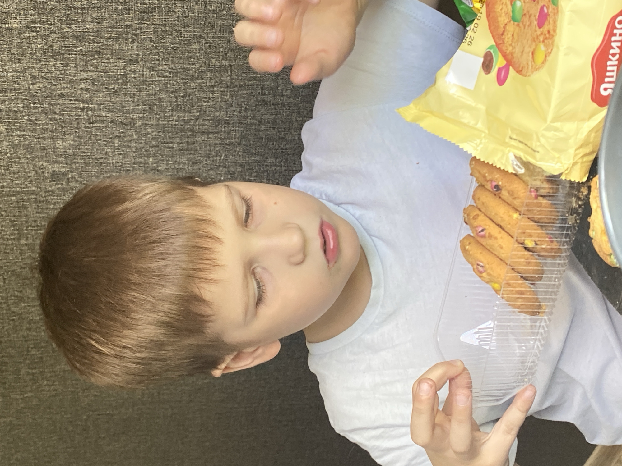 A child lies on a gray surface, possibly carpet. They are fair-skinned with light brown hair and are wearing a white t-shirt. Their eyes are closed, and their mouth is slightly open. A clear plastic container of cookies sits beside them, along with a yellow package labeled "Яшкино". The location and city cannot be determined from the image alone.