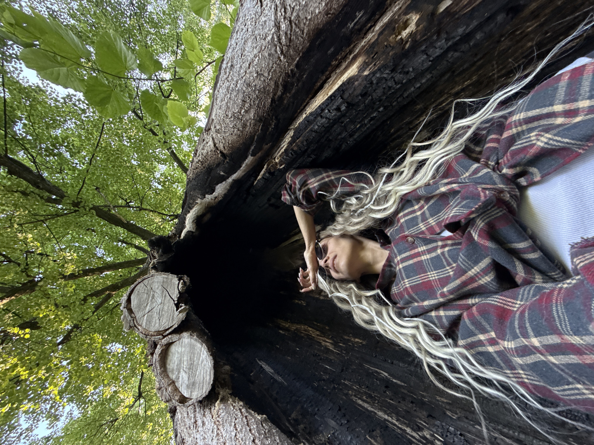 A low-angle photograph, rotated 90 degrees to the left, captures a person resting inside a large, hollow tree trunk within a wooded area, likely in Rusanovo, Russia. The tree trunk appears to run diagonally upwards, framing the scene.
The main subject is a person with long, wavy, blonde hair, wearing a red, black, and beige plaid flannel shirt over a light-colored garment. They are lying on their back within the dark, hollow interior of the tree trunk, looking upwards. Sunglasses are visible, resting on their head or hair, and their right hand is gently placed near their face, conveying a sense of relaxation or contemplation.
The hollow interior of the tree trunk is very dark, appearing charred or deeply decayed, with a rough, textured surface. The exterior bark, visible on the edges of the trunk, shows natural wood tones. On the left side, two circular cross-sections of cut branches are visible, displaying concentric wood grain.
Above and beyond the tree trunk, a vibrant canopy of bright green deciduous leaves fills the background against a light sky. This dense foliage indicates a forest or heavily wooded setting. The bright, natural light suggests it is daytime, likely morning or afternoon, under clear weather conditions. There is no visible text in the image.