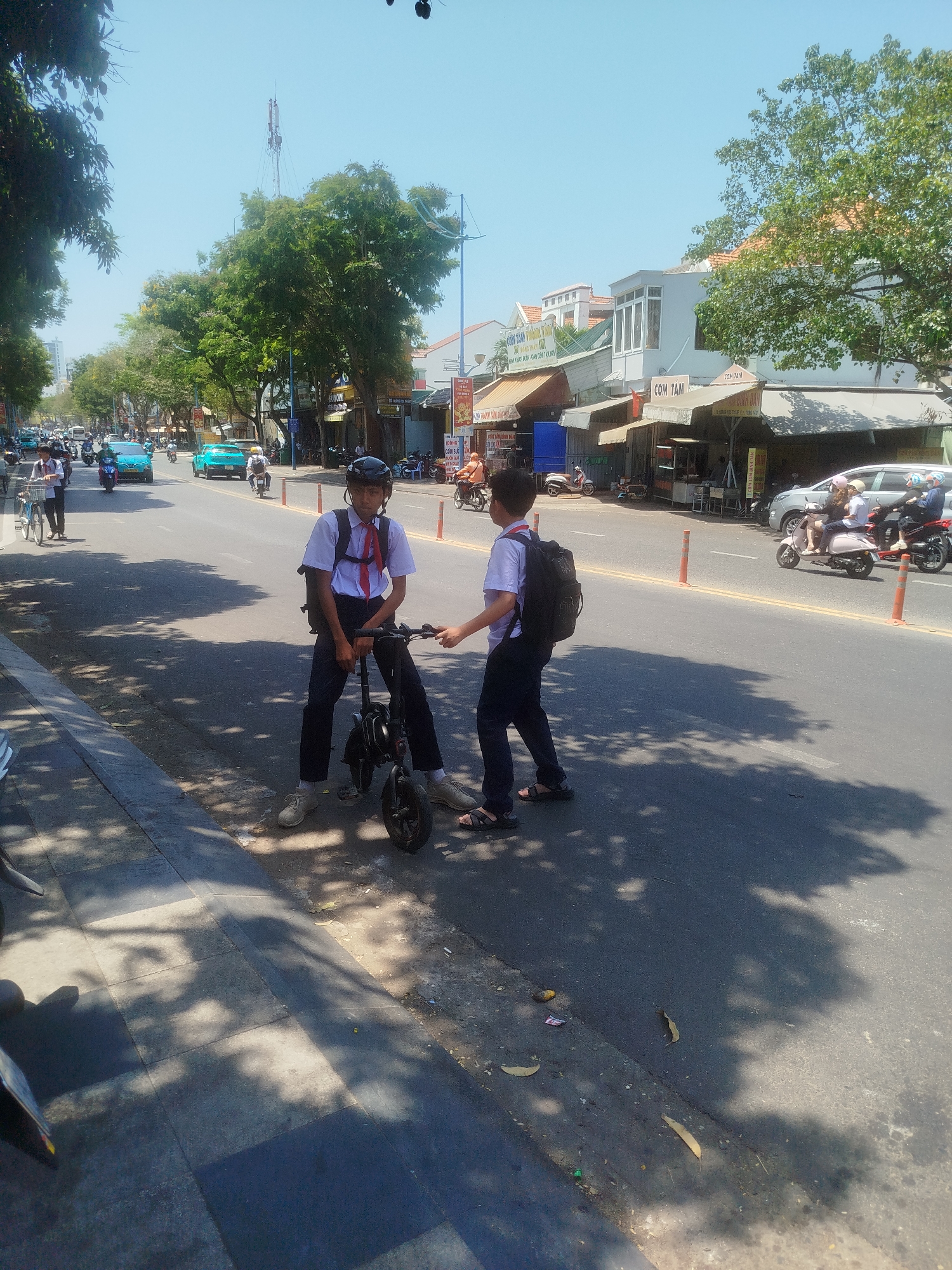 Two schoolboys in uniform stand on a road in Ho Chi Minh City, Vietnam, next to a small, foldable electric scooter. One boy is wearing a helmet and is sitting on the scooter, while the other stands beside him with a backpack. They are positioned on a sunny street lined with shops and trees. Traffic, including motorbikes and cars, passes by in the background.