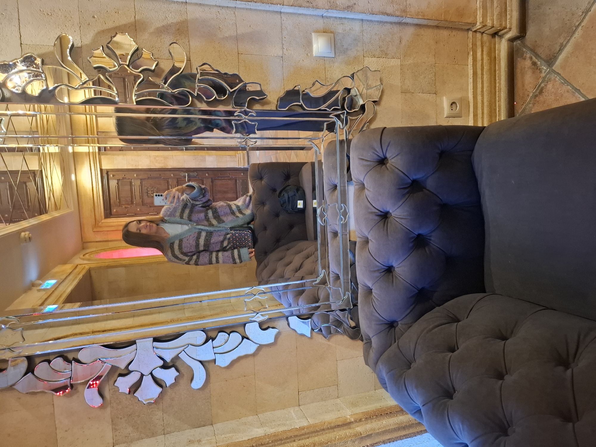 A medium shot captures a gray, tufted couch in a room with a beige, textured wall. A woman, reflected in an ornate mirror, stands in the background holding a mobile phone. The mirror's decorative trim is composed of silver, cut-glass elements. The location appears to be in Zadonsk, Russia.
