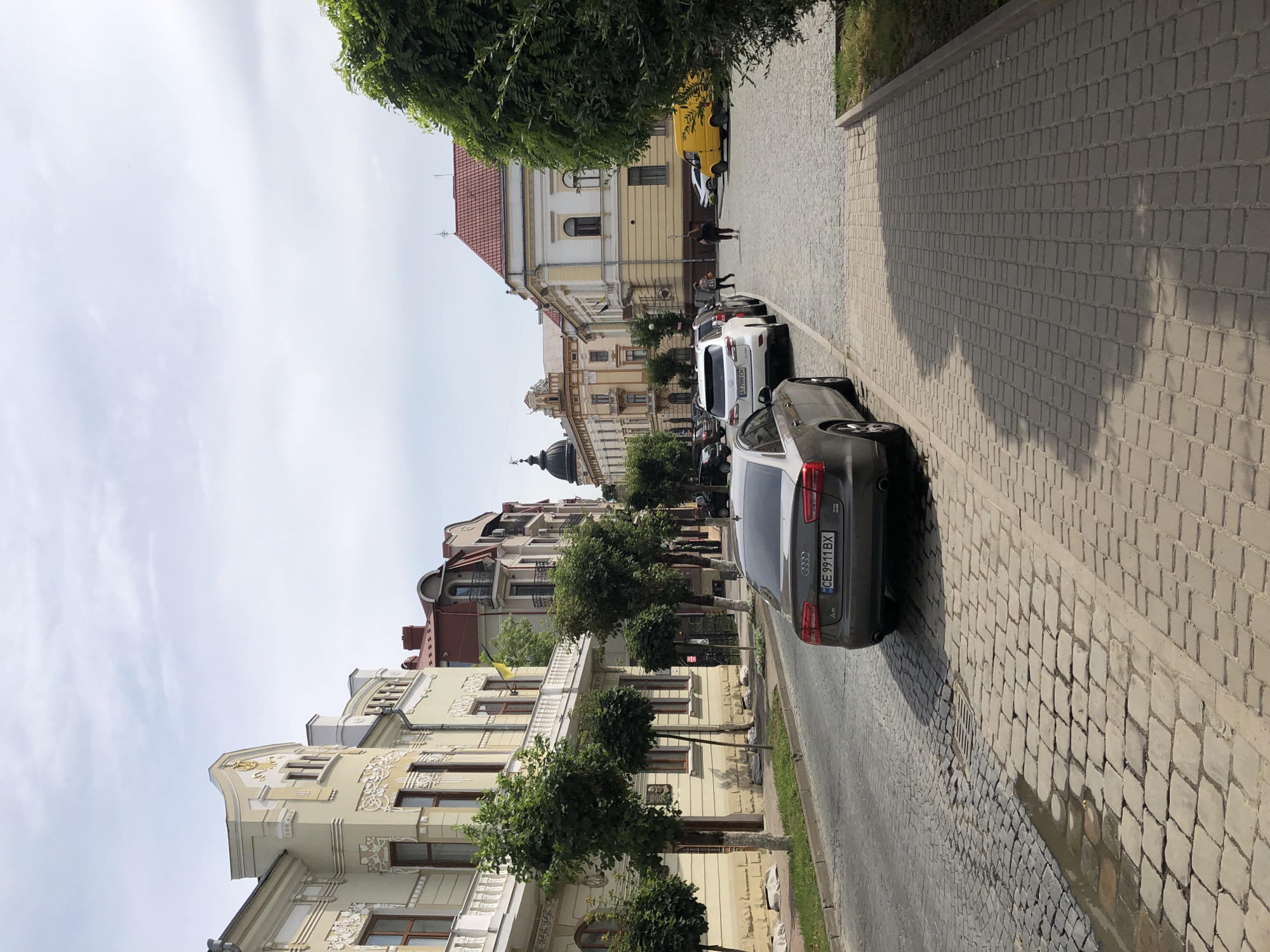 The image depicts a street scene in Kolomyia, Ukraine. The street is paved with cobblestones. On the left side of the street, there are several buildings with ornate architecture, featuring balconies and detailed facades. Small, neatly trimmed trees are planted along the sidewalk in front of these buildings.
In the foreground and midground, several cars are parked along the street. A dark grey Audi is prominently featured in the foreground, facing away from the camera, with its license plate visible. Other cars, including a white Lexus and a white sedan, are parked further down the street.
On the right side of the street, a paved sidewalk runs alongside a green, leafy tree. Beyond the sidewalk, more buildings are visible, including a building with a red tile roof. A yellow van is parked in the distance.
A few people are walking on the sidewalk in the middle distance. The sky is overcast with scattered clouds. The overall impression is of a quiet, well-maintained urban street in a European city.