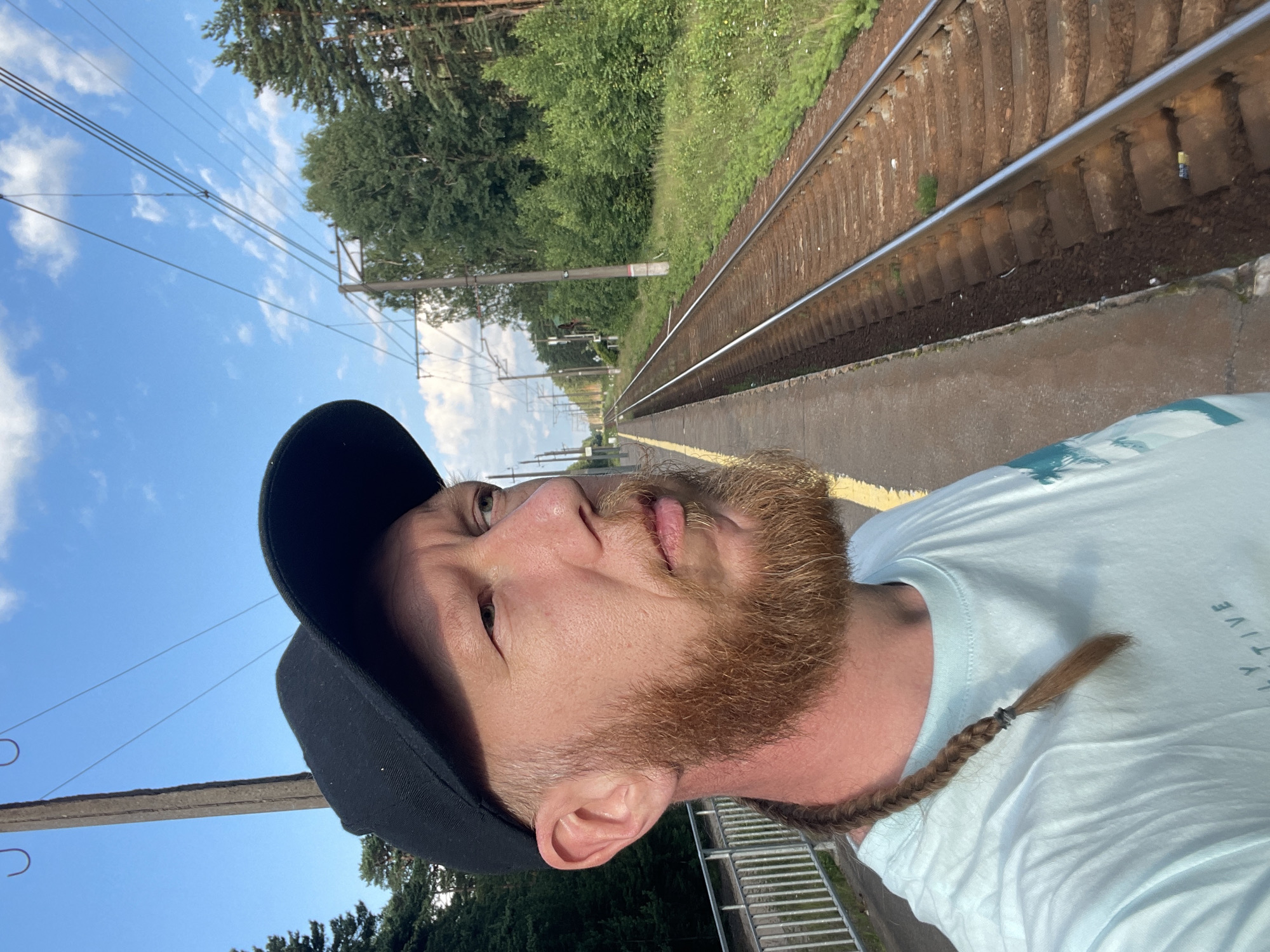 A man with a braided beard and a black baseball cap stands on a train platform in Lebyazhye, Russia. He is wearing a light blue t-shirt. To his right, train tracks stretch into the distance, flanked by trees and vegetation. The sky is blue with scattered clouds. Overhead, power lines are visible. A metal fence runs along the edge of the platform.