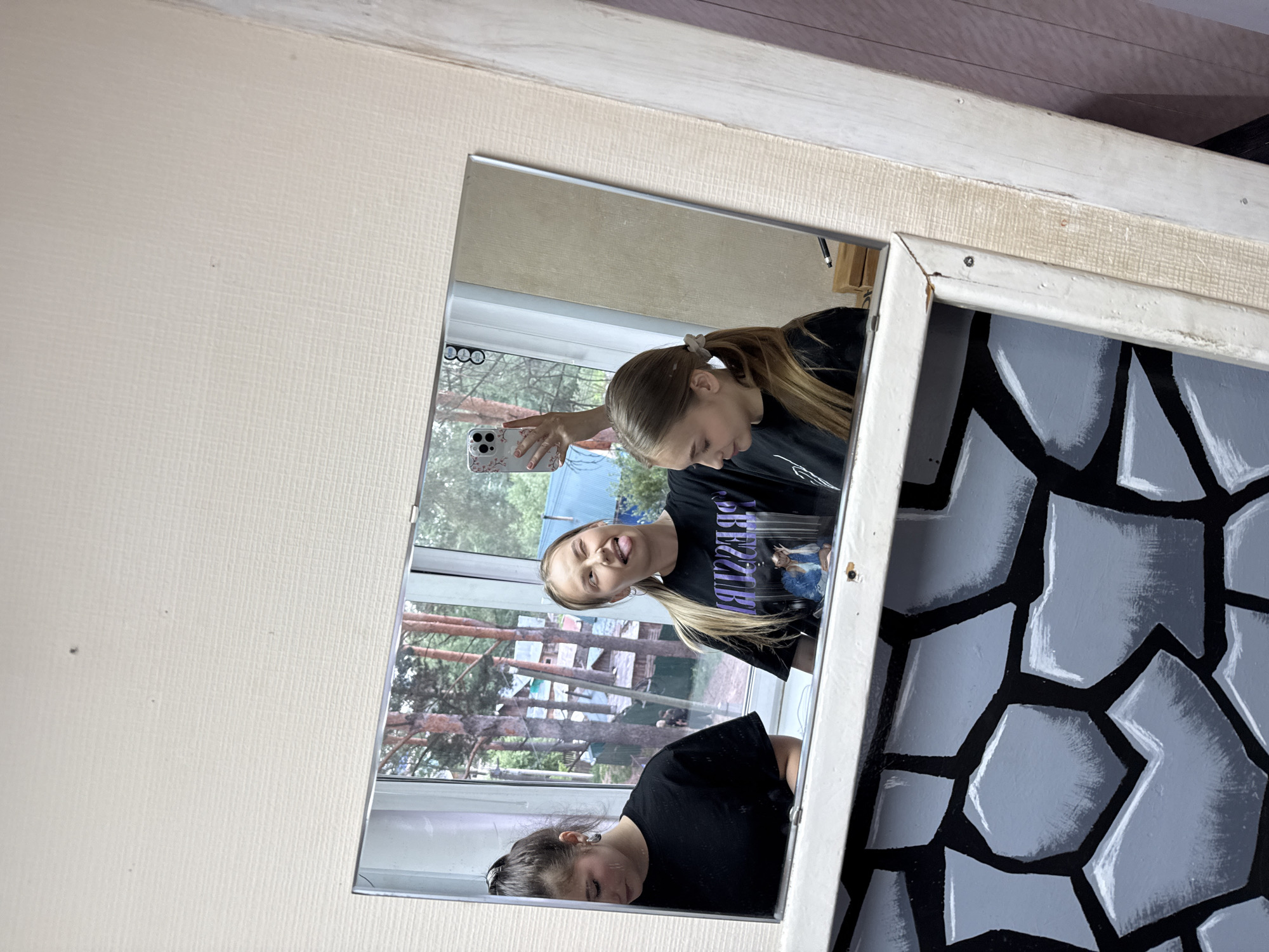 Two girls are reflected in a mirror. One girl is holding a phone and taking a selfie. The reflection shows a view from a window with trees outside. The mirror is decorated with a pattern resembling stones. This is likely located in Chaikovsky, Russia.