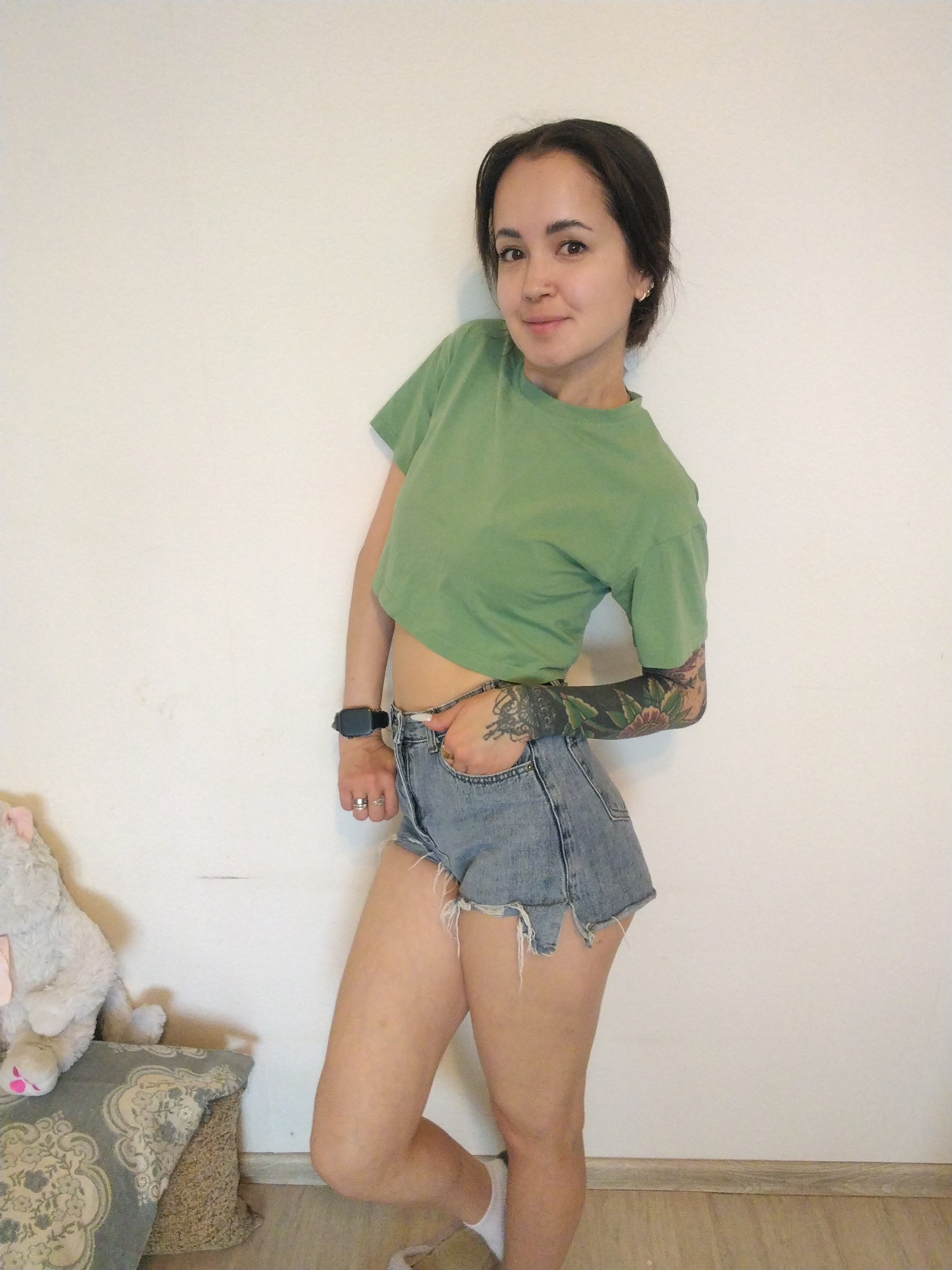 A woman stands indoors against a plain white wall. She has dark hair and fair skin. She wears a short-sleeved green shirt, denim shorts, a wristwatch on her left wrist, and white socks. Her left arm is extensively tattooed. A gray stuffed animal and a patterned pillow are to her left on the floor. The floor is a light wood. The location is not apparent from the image. There is no discernible evidence of a city or the specific location of Noviy, Russia.
