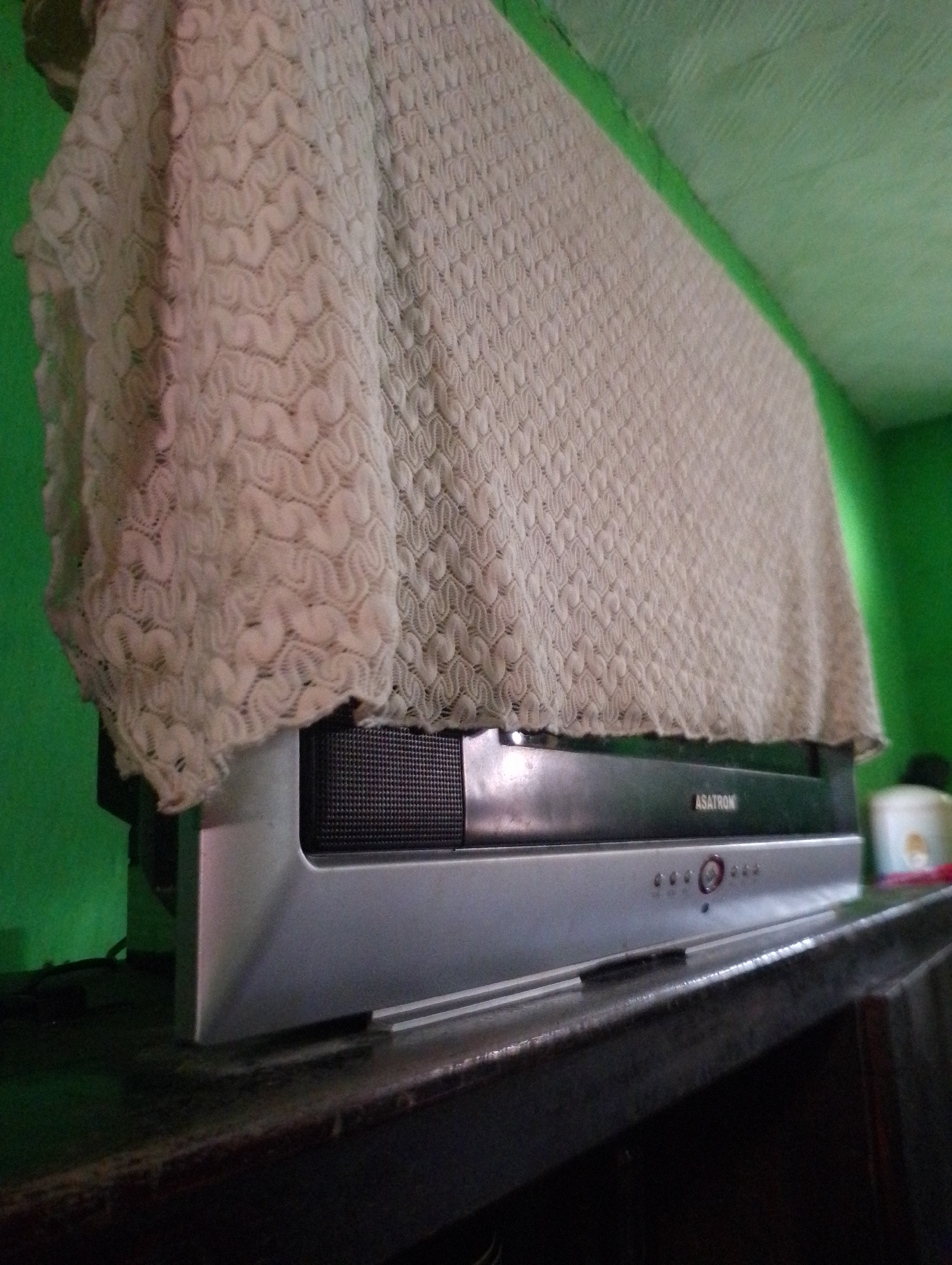 A television set is partially covered by a cream-colored, textured fabric. The television appears to be a flat-screen model with a silver bezel and a black speaker grille. The brand name "ASATRON" is visible on the front of the television. The television is positioned on a dark wooden surface, likely a table or shelf, in front of a bright green wall. The fabric cover has a repeating pattern of interconnected shapes. The lighting in the image is somewhat dim, casting shadows. The overall scene suggests an indoor setting, potentially a home. The context provided indicates this location is in Sukamanah, Indonesia.