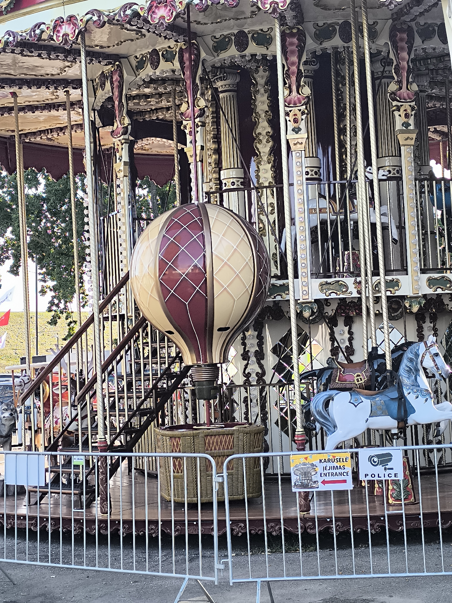 The image shows a carousel with a hot air balloon figure hanging prominently in the foreground. The carousel is elaborately decorated with ornate columns, gold trim, and detailed carvings. Several carousel horses are visible, including a white and blue horse in the mid-ground. In the background, trees and a grassy embankment can be seen. A metal fence encloses the carousel area, with signs indicating "KARUSELIS IEJIMAS ENTRANCE" and "POLICE" with a security camera icon. The carousel appears to be set up outdoors. The overall impression is of a festive or amusement park setting.