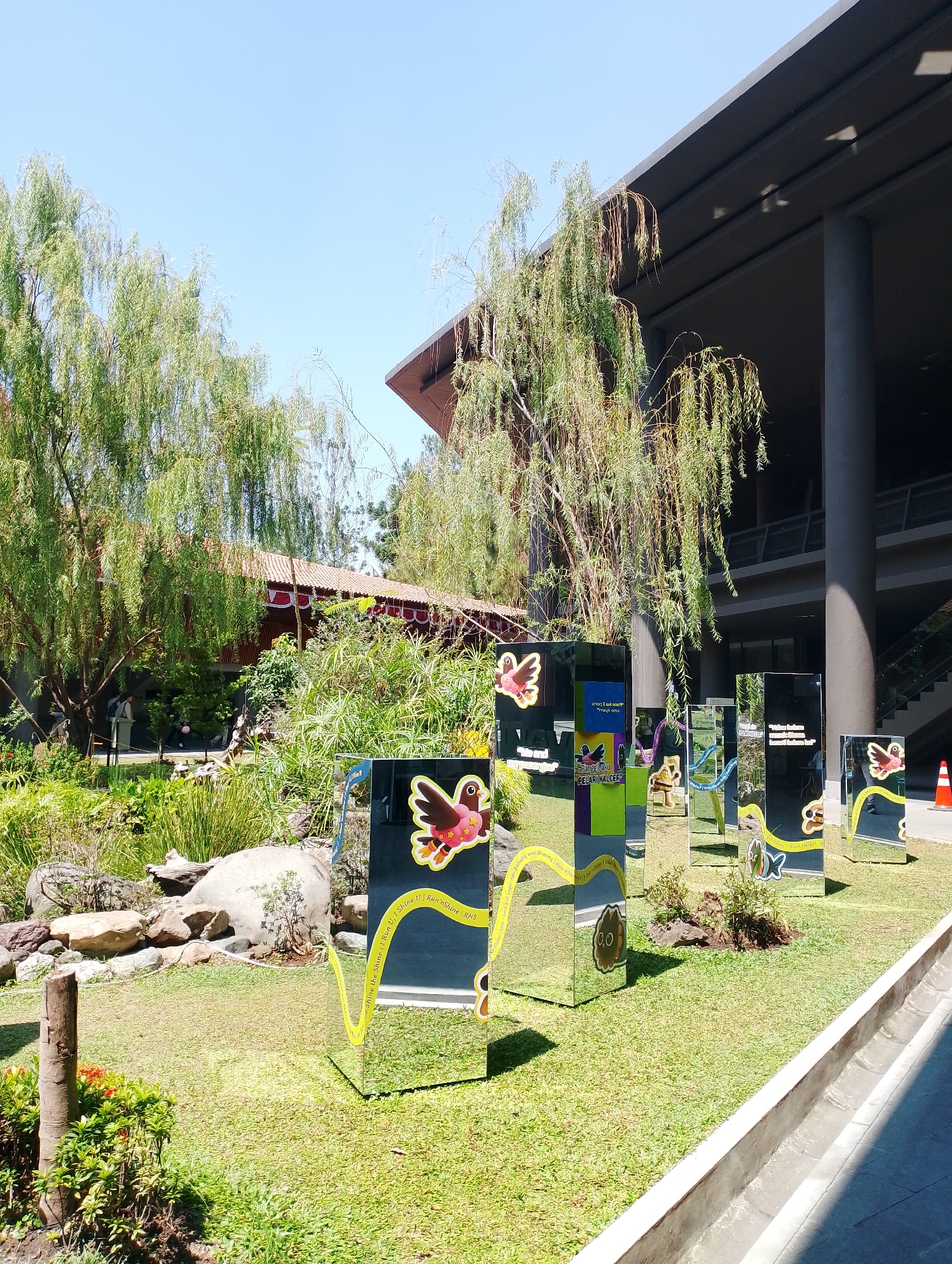 This image shows an outdoor exhibit featuring mirrored pillars decorated with colorful cartoon bird graphics and text. The pillars are arranged on a grassy area in front of a building with a dark, modern architectural style, and a traditional building with a red tiled roof. Lush green trees and dense vegetation surround the exhibit. In the foreground, there is a small pond with rocks and plants. The sky is clear and blue. The location is Depok, Indonesia, specifically at the Bird Park within the Taman Mini Indonesia Indah.
