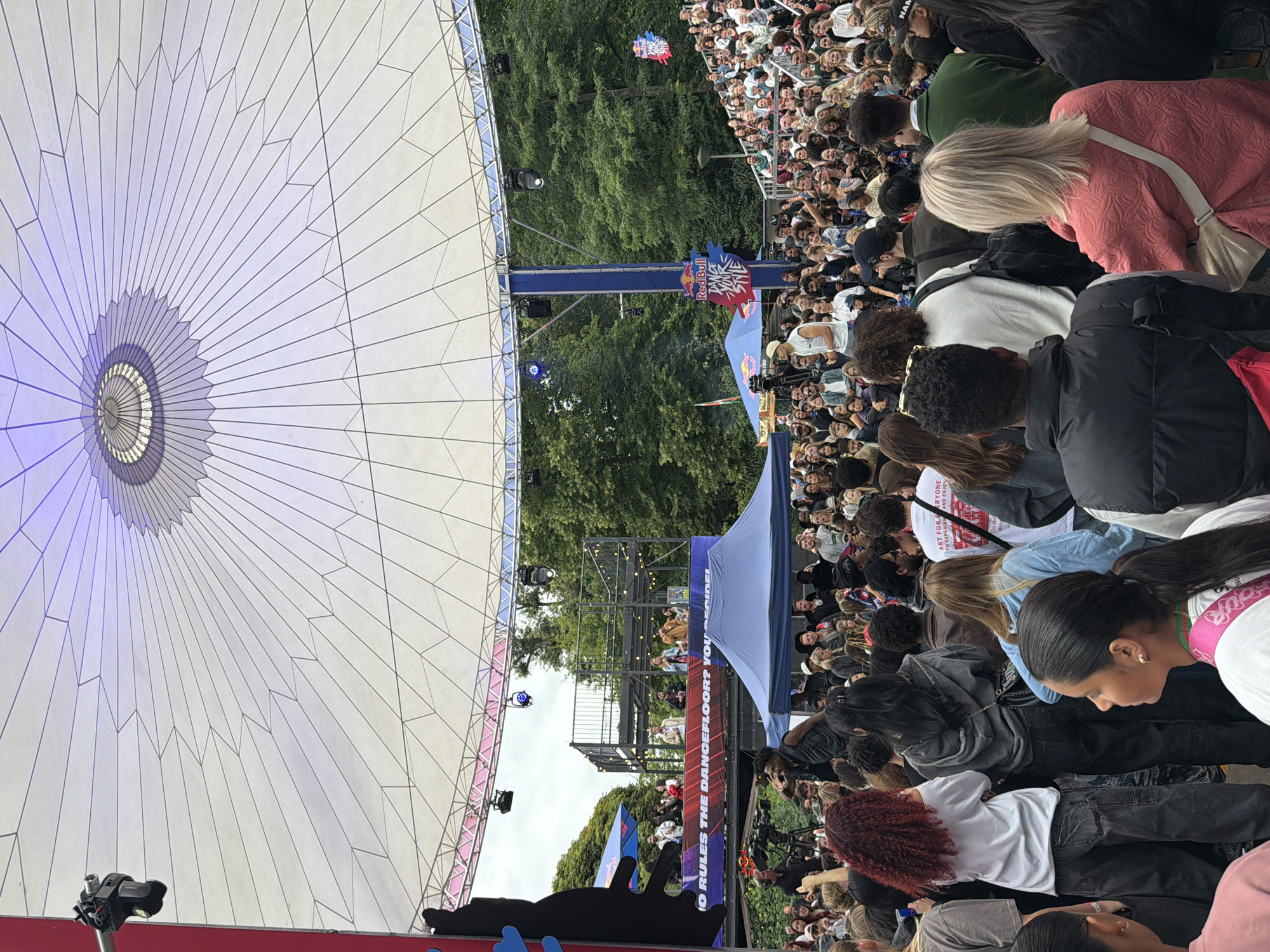 This media file depicts an outdoor event, likely a dance competition or performance, held in Amsterdam. A large crowd of spectators is gathered, many of whom appear to be young adults. They are positioned in front of a stage structure, partially covered by a large white canopy. The canopy's design is reminiscent of a sunburst, with a central circular element radiating outward into many triangular panels.
Behind the stage, a dense line of trees suggests a park or wooded area. The lighting indicates it's daytime, though the sky is not prominently visible. Several individuals are on a raised platform on the stage, possibly performers or judges. The atmosphere seems energetic and engaged, with people leaning forward and looking towards the stage.
Prominent text visible in the image includes "Red Bull," a sponsor logo, and "O RULES THE DANCEFLOOR? YOU," implying a competitive or rule-based dance event. Other text, "ART FOR EVERYONE TO EXPERIEN AND ENJOY," suggests a broader artistic or community focus. The presence of cameras and lighting equipment indicates a professionally organized event. The overall impression is one of a vibrant, public gathering centered around a dynamic performance or competition, with strong branding from Red Bull.