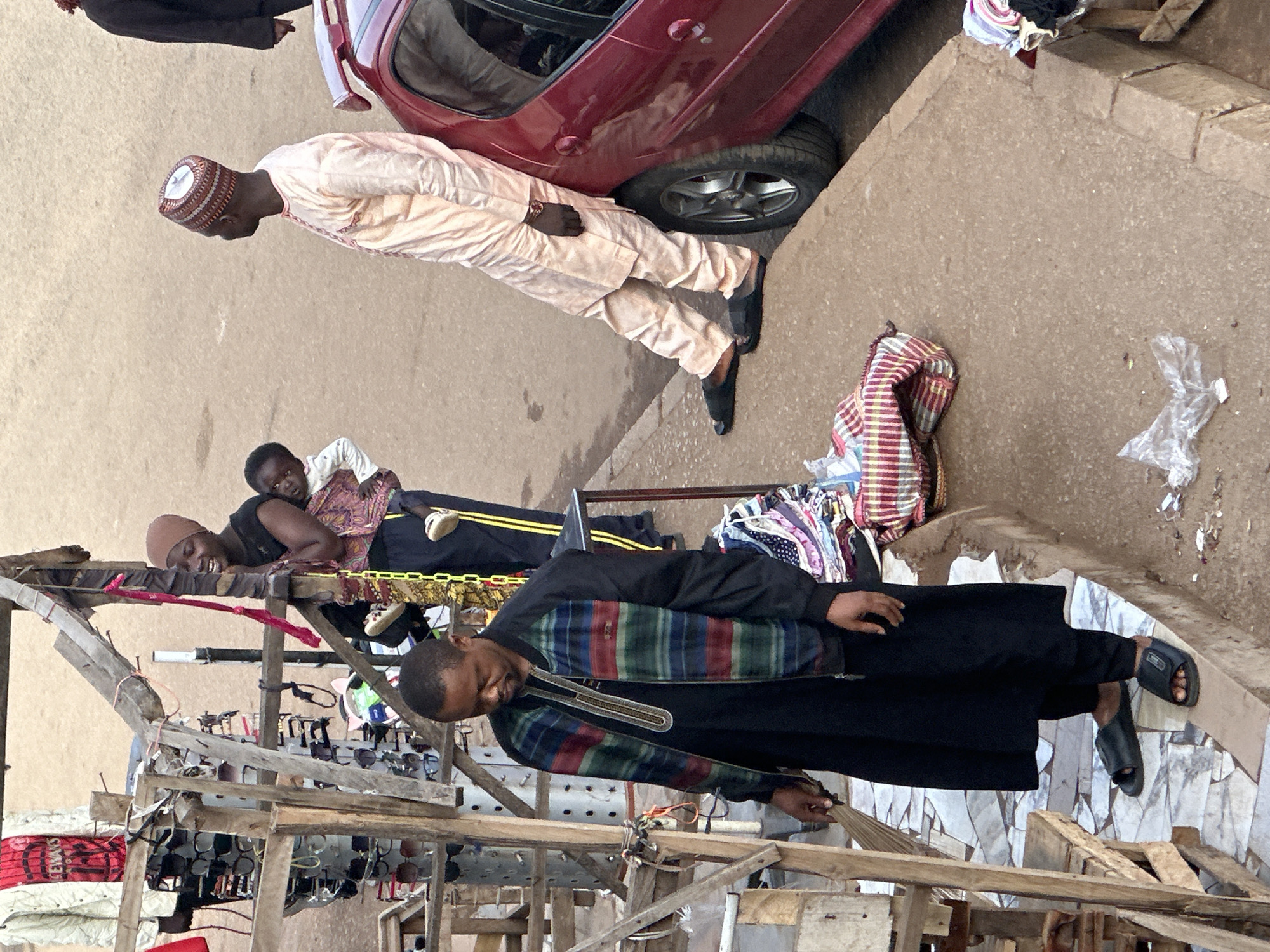A street scene in Bukuru, Nigeria, features a man in a black robe and a colorful striped jacket, who appears to be sweeping or attending to a stall with wooden structures displaying sunglasses. He is wearing black sandals. To his right, a woman wearing a headscarf and black top is holding a baby. Further to the right, a man in a light-colored traditional outfit and a patterned cap leans against a red car. The ground is a mix of pavement and dirt. Several pieces of fabric and plastic are scattered on the ground. The overall scene depicts a bustling marketplace environment.