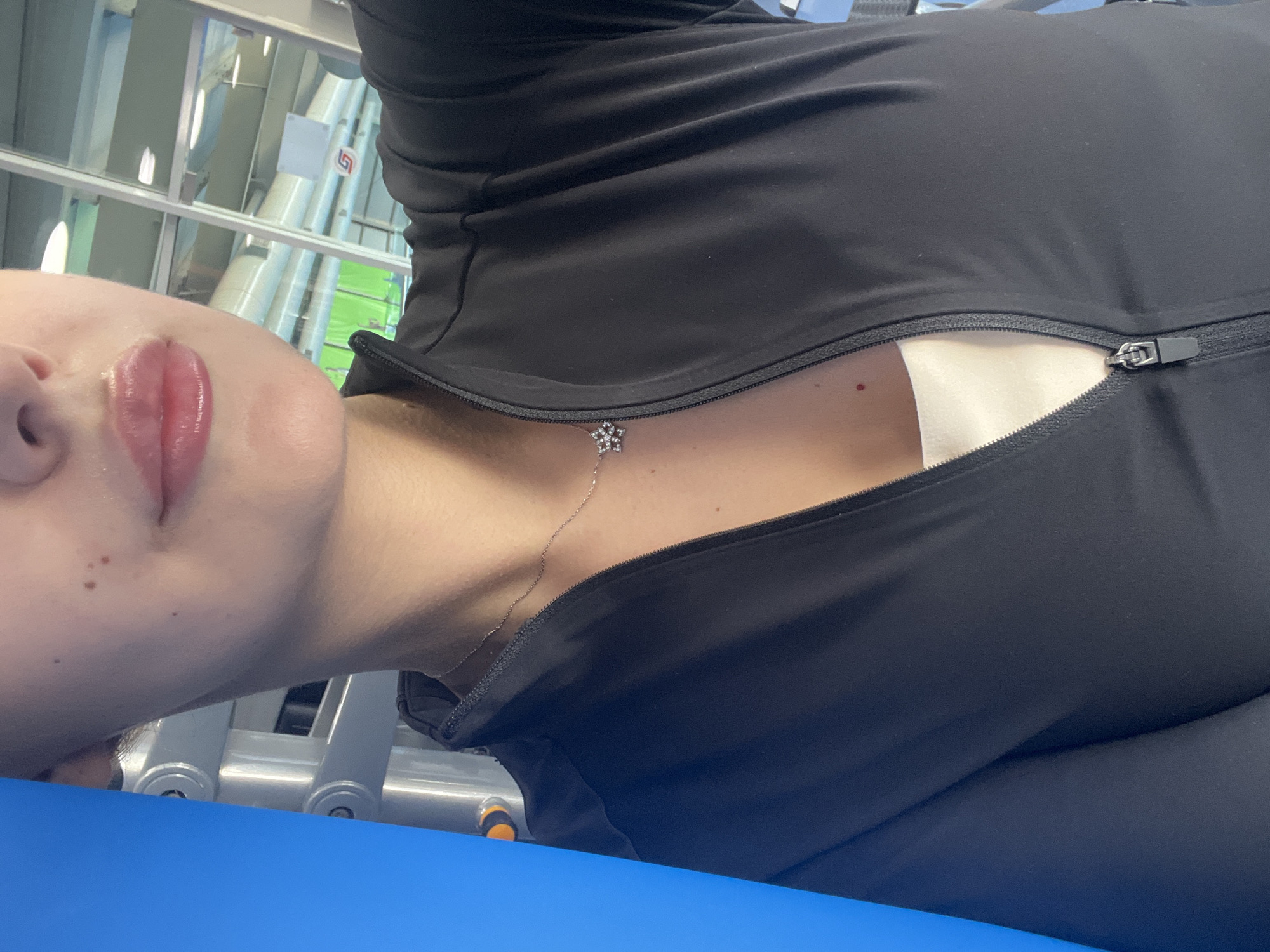 A close-up, slightly angled, eye-level shot captures a person lying on a blue surface, possibly a gym bench or exercise mat. The individual is wearing a black zippered athletic top, which is partially unzipped revealing a white undergarment. A delicate silver necklace with a small star-shaped pendant rests on their chest. The person's face, partially visible, shows full pink lips.
In the blurred background, what appear to be metal structures and windows suggest an indoor setting, possibly a gym or fitness center in Yerevan, Armenia. The environment includes linear patterns, which might be structural supports of the building.