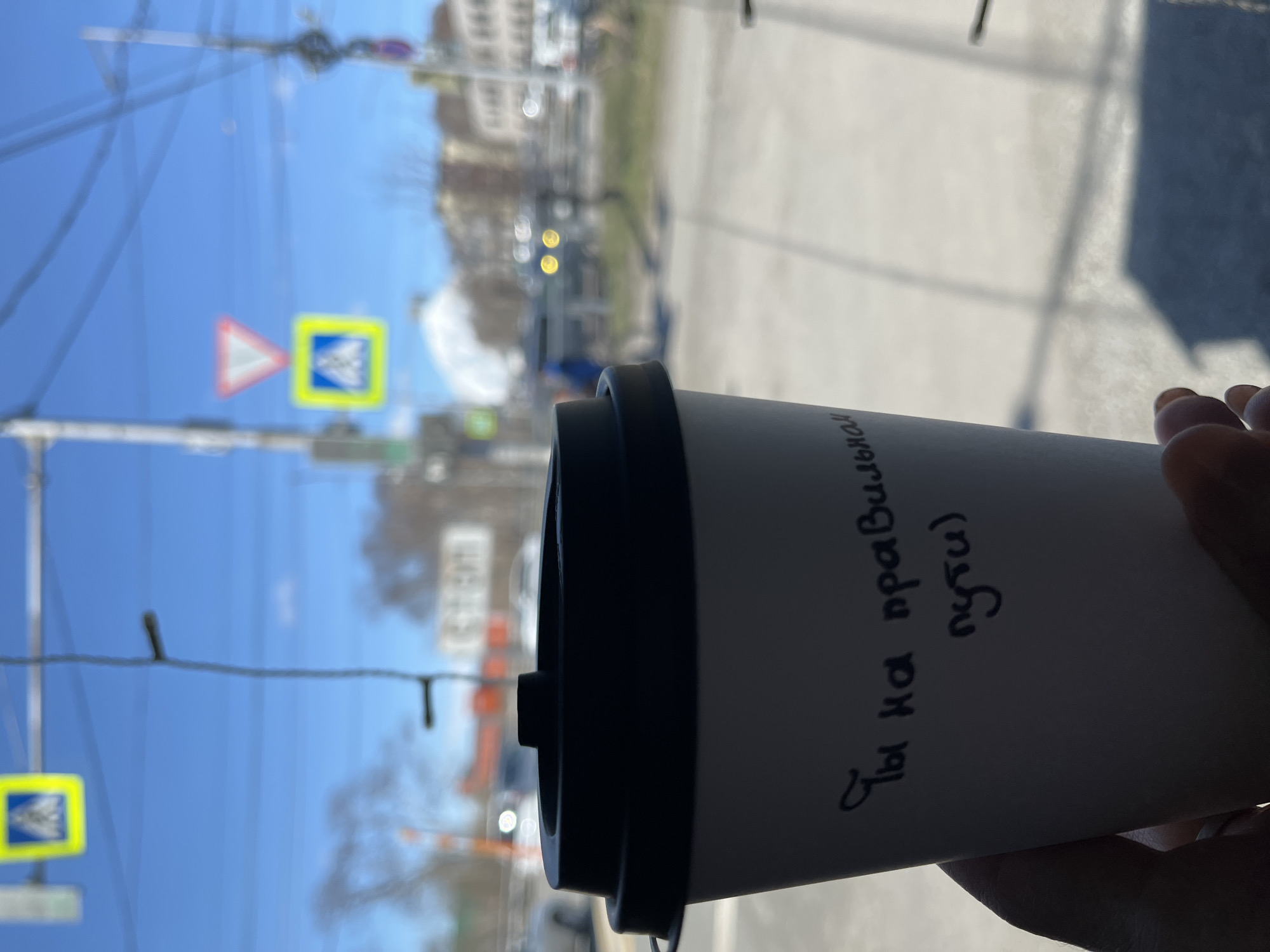 A person is holding a coffee cup with black lid in front of a window, with a street scene of Petropavlovsk-Kamchatsky, Russia, visible through the glass. The text “Ты на правильном пути)” is handwritten on the cup. Outside the window there are: blue skies, buildings, a pedestrian crossing sign, power lines, and vehicles. The street appears to be a roadway with vehicular and pedestrian traffic.