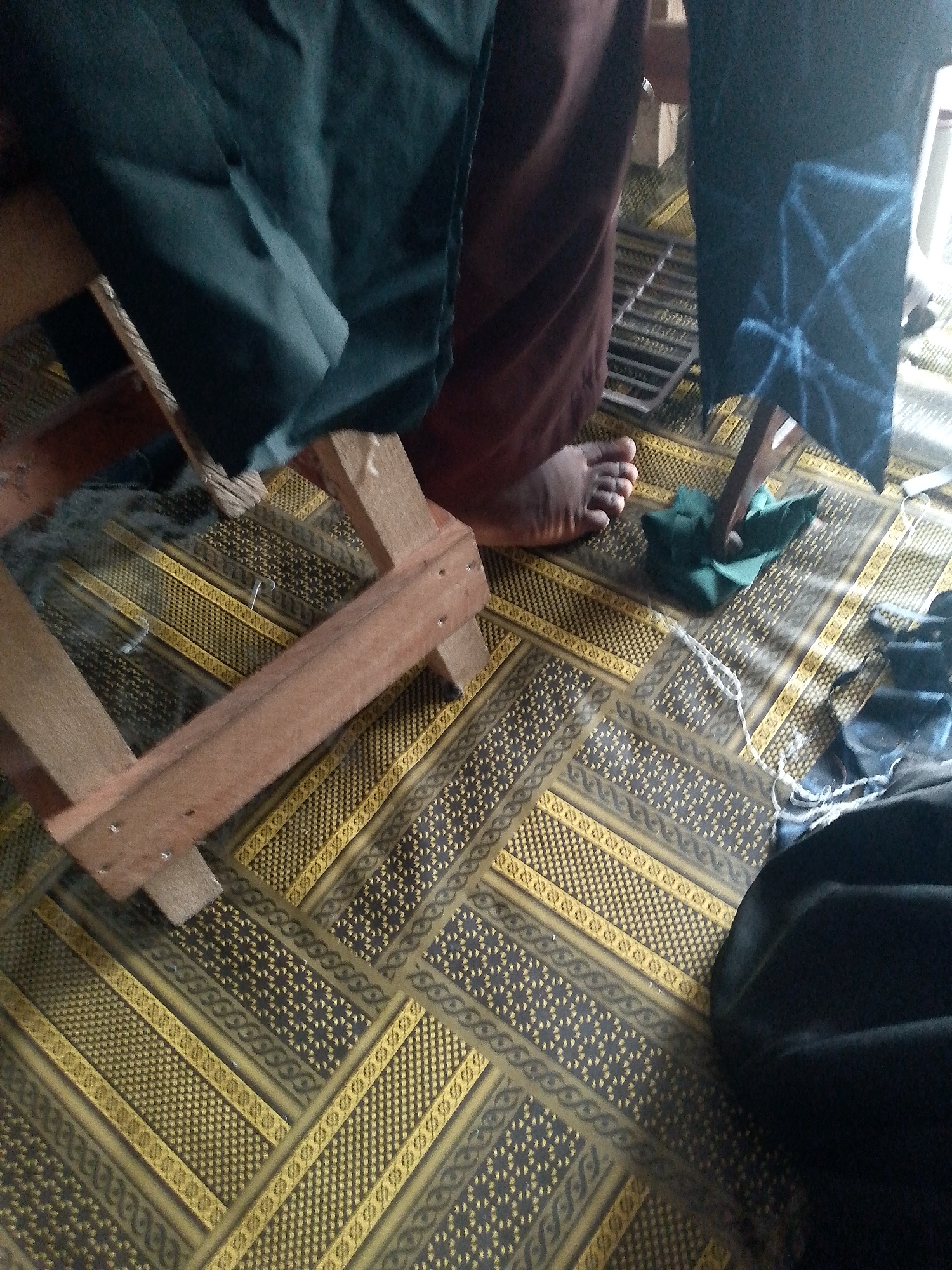 The image depicts a close-up view of a person's foot and legs in a room with a patterned floor. The floor is covered with a mat or rug featuring a repeating geometric design in yellow, black, and dark grey. A wooden structure, possibly part of a loom or furniture, is visible, along with pieces of dark fabric. One piece of fabric has a white geometric pattern. A small crumpled piece of green fabric is on the floor near the wooden structure. The scene appears to be indoors, given the lighting and the presence of fabric and a floor covering. The context of Jauro Yinu, Nigeria, suggests a cultural or craft setting where traditional textiles might be produced or displayed.