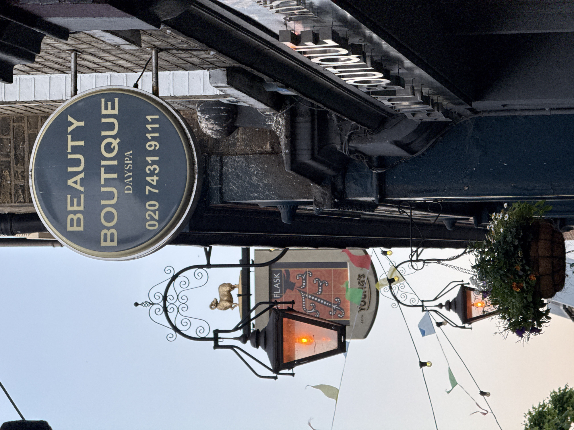 The image shows an exterior scene in London, United Kingdom, featuring a "Beauty Boutique" sign displaying the phone number "020 7431 9111". Below, there's a "Flask" sign indicating a pub, complete with a decorative wrought iron lantern with an illuminated bulb. A hanging basket of flowers and string lights with small flags are visible in the background. The overall composition is viewed from a low angle, looking upwards.
