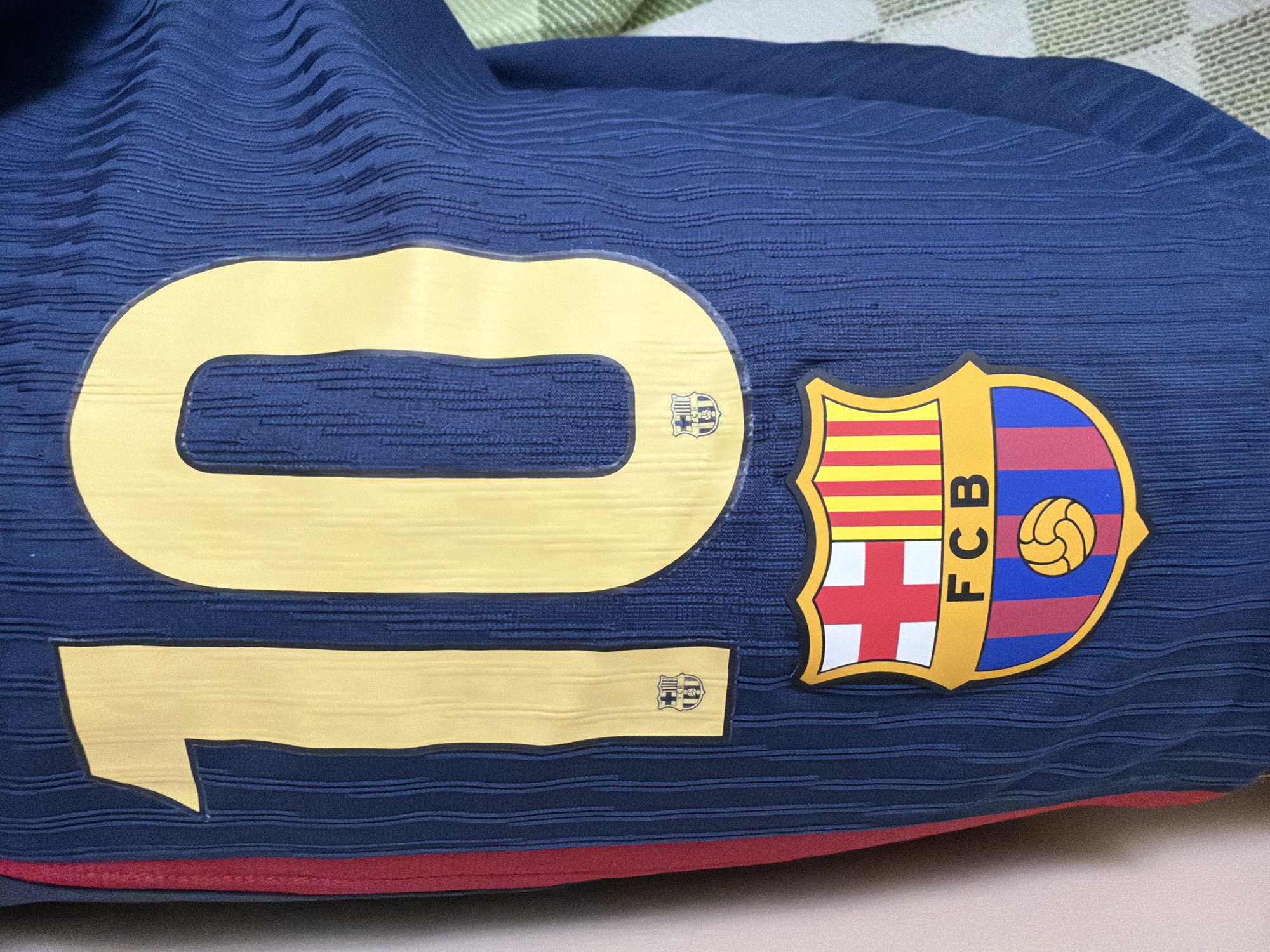 The image shows a close-up view of a blue soccer jersey with yellow numbers "10" on the front. The jersey features the crest of FC Barcelona, a shield with red and white quarters, yellow and red stripes, and a blue and red lower section containing a yellow soccer ball. There are also two smaller FC Barcelona crests on the yellow "10". The jersey appears to be made of a textured fabric. The background is out of focus and shows a light-colored surface to the left and a checkered pattern to the right.