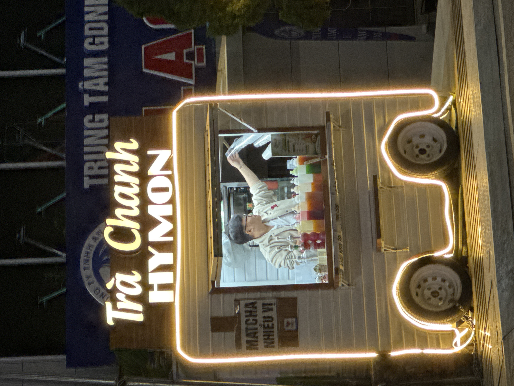 A brightly lit food truck with neon lights and the words "Trà Chanh HYMON" is parked on a street in Da Lat, Vietnam. A young man wearing glasses and a light-colored jacket is inside the truck, preparing drinks. He is reaching for a plastic cup and appears to be serving a customer. Various colorful syrups and cups are visible on the counter inside the truck. A sign on the left side of the truck reads "MATCHA NHIỀU VỊ", indicating that matcha drinks with multiple flavors are offered. The food truck has two wheels visible, and the surrounding area is dimly lit, suggesting it is nighttime. A large blue banner with Vietnamese text is visible in the background, partially obscured by the food truck.