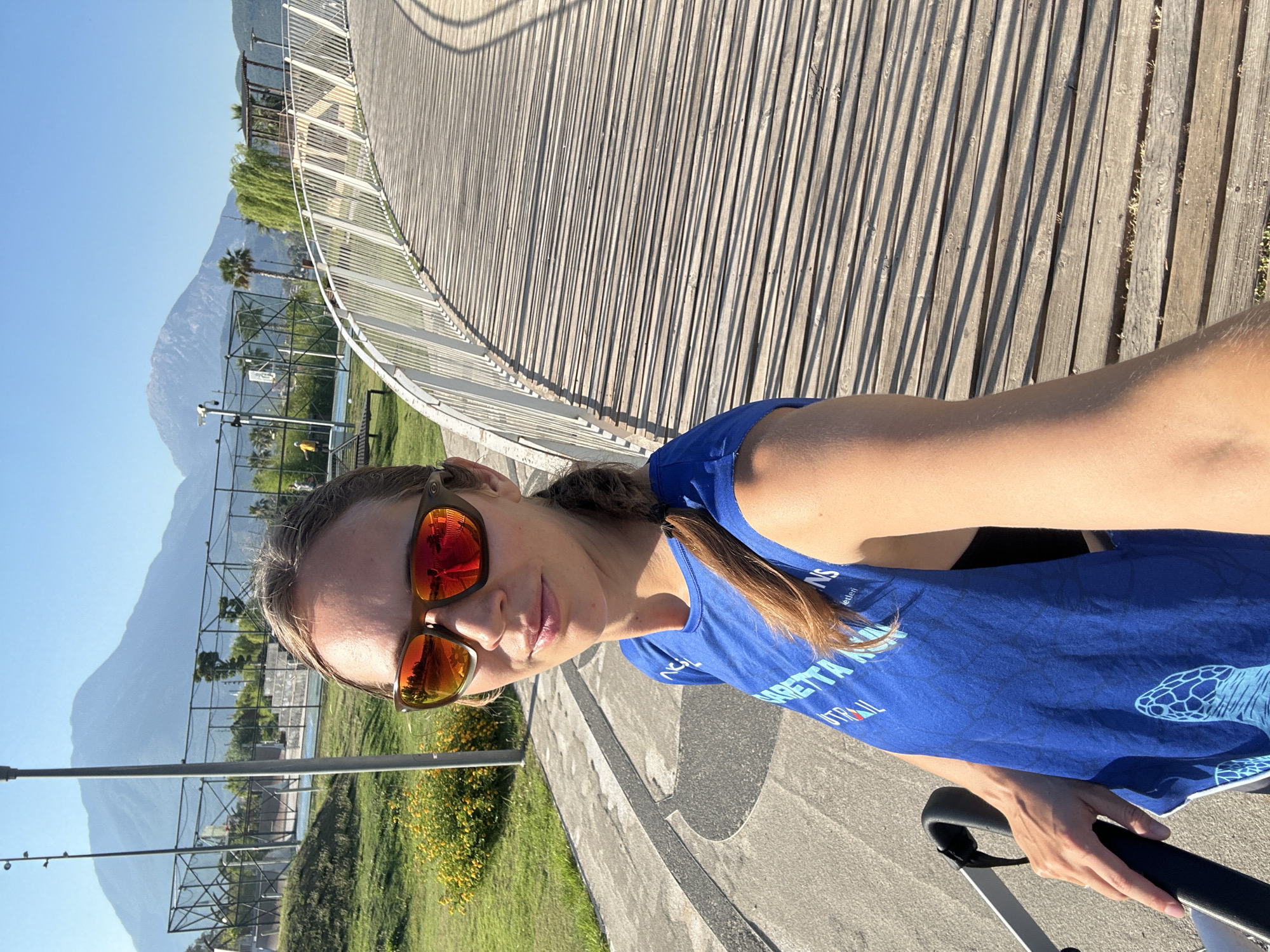 A woman is taking a selfie outdoors, with a wooden boardwalk curving behind her. She is wearing sunglasses with orange lenses that reflect the bright sunlight, and a blue t-shirt with some text and a graphic of a turtle on it. Her hair is pulled back into a ponytail. The scene appears to be a park or recreational area, with green grass, yellow flowers, and trees visible in the background. In the distance, mountains rise against a clear blue sky, and there is a sports court with a fence. The time of day appears to be morning or late afternoon, judging by the long shadows cast by the boardwalk railings. The overall mood seems relaxed and active, suggesting the woman might be enjoying a run, walk, or bike ride. Text on her shirt appears to include "SETTA" and "TRAIL". The location is Fethiye, Turkey.