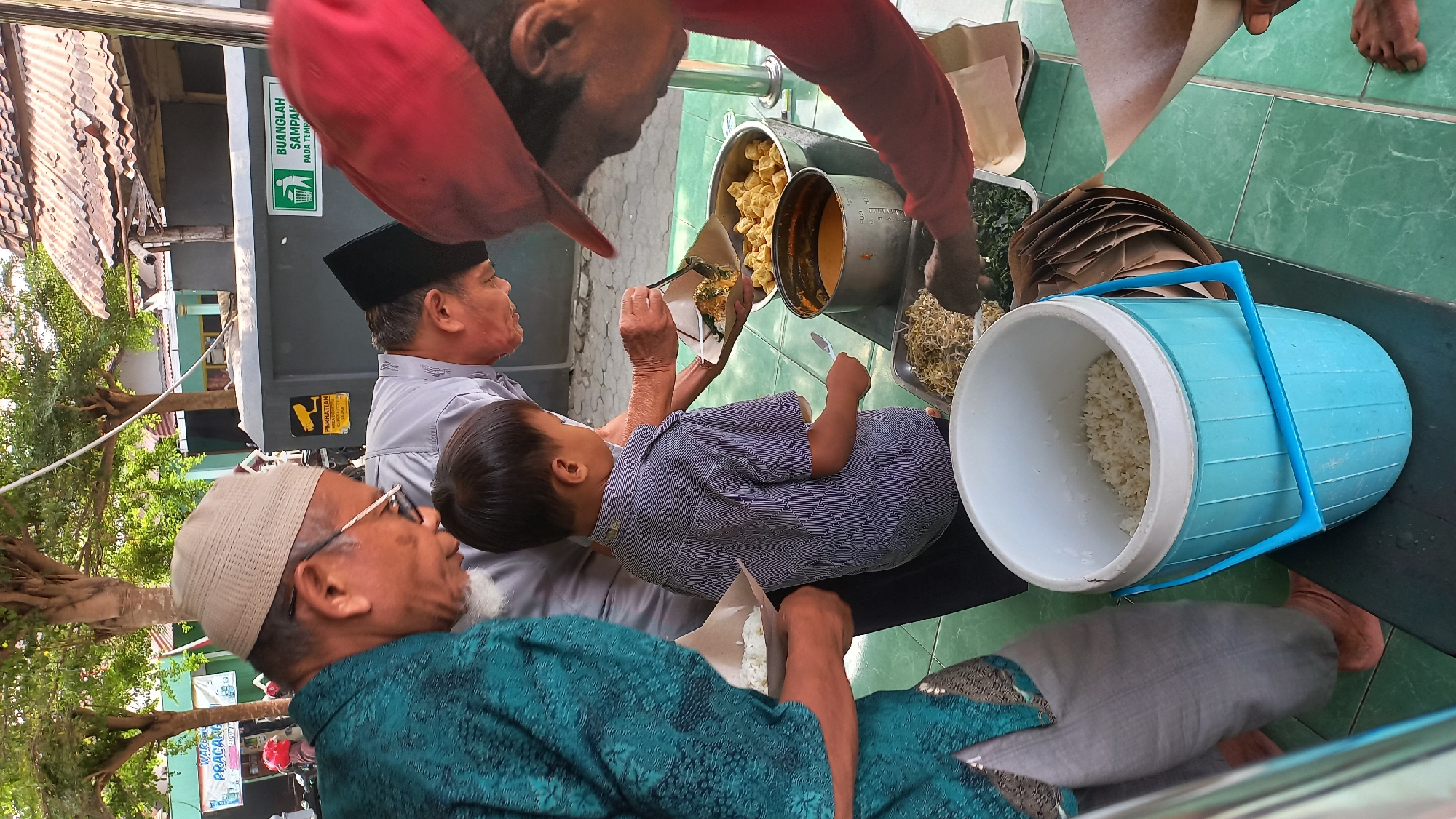 In Kenayan, Indonesia, an outdoor food distribution event is underway on a green tiled surface. Several individuals are gathered around a table laden with food. In the foreground, an elderly man wearing a teal patterned shirt and a beige cap is holding a cone of white rice. Beside him, a young boy in a striped shirt stands holding a spoon. To the right, a person wearing a red cap is serving food onto a cone of paper for another individual.
On the table, there are several large metal containers holding various dishes, including what appears to be cooked vegetables, bean sprouts, and a saucy mixture. A large, light blue insulated cooler filled with white rice is also present. In the background, there are buildings with tiled roofs and trees, suggesting a public or community setting. Signs are visible on a wall, one with a CCTV camera symbol and another with a trash can icon and text in Indonesian. The overall scene depicts a communal meal being served.