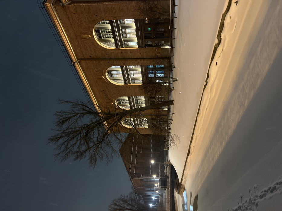 A brick building with large arched windows stands next to a snowy landscape under a dark sky. Trees with bare branches are visible near the building. Street lights illuminate a path along the base of another brick building further back. The photo is angled so that the building and the street lights run vertically and the sky horizontally.