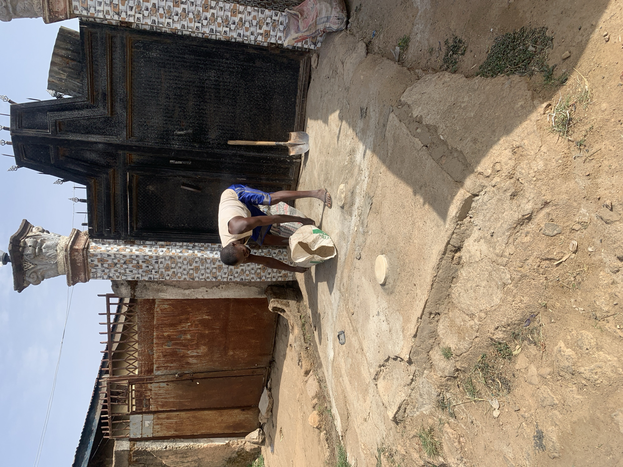This outdoor image, taken in Jos, Nigeria, captures a scene of manual labor under a clear blue sky, indicating bright daylight. Strong shadows cast on the ground suggest it is mid-morning or afternoon.
The central figure is a dark-skinned man, dressed in a light-colored short-sleeved shirt and blue shorts. He is partially barefoot, with one sandal on his right foot. He is bent over, intently focused on a white sack on the ground, appearing to be working with its contents, possibly mixing or preparing materials.
The setting features a blend of concrete pathways and dry, earthy ground with sparse patches of grass. Dominating the background is a large, dark, ornate metal gate with decorative spikes along its top, propped open. A shovel leans against this gate. Adjacent to it, a wall adorned with small, patterned mosaic tiles in shades of brown, white, and black is visible, alongside a decorative concrete pillar with intricate carvings. To the far left, a smaller, rusty, corrugated metal gate can be seen.
Additional objects include a larger, partially visible bag leaning against the tiled wall, likely containing building materials. A small, round, light-colored object rests on the concrete near the man. The general appearance of the architecture, the presence of construction materials, and the man's activity suggest an area undergoing maintenance or development. No distinct text is clearly readable on the sacks.