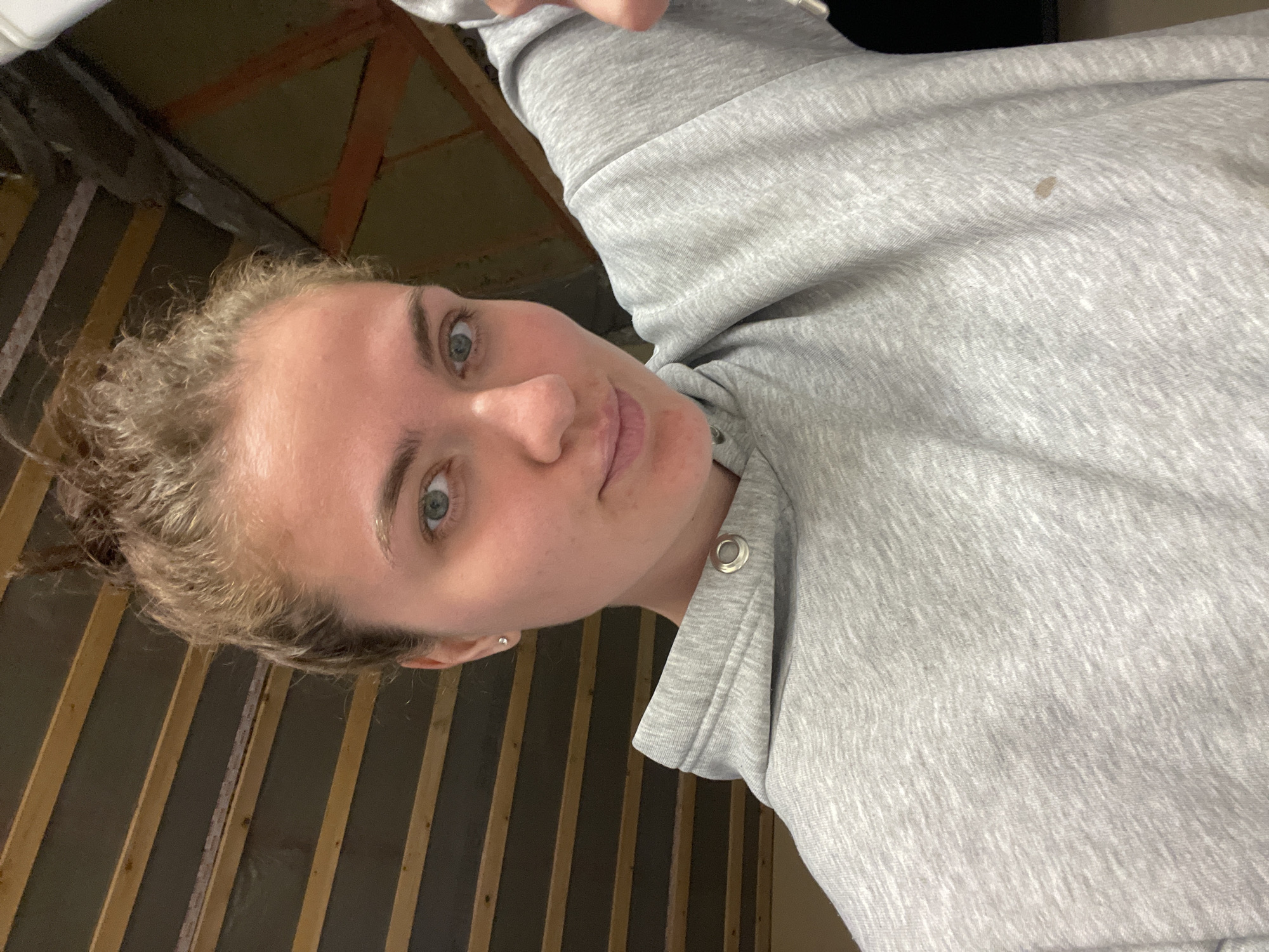 A woman with fair skin and light, curly hair, tied back, is looking directly at the camera. She has blue eyes and a neutral expression. She is wearing a light grey hooded sweatshirt. The background consists of dark vertical planks and structural beams. The location is Мосметаллоконструкция Онт, Russia.